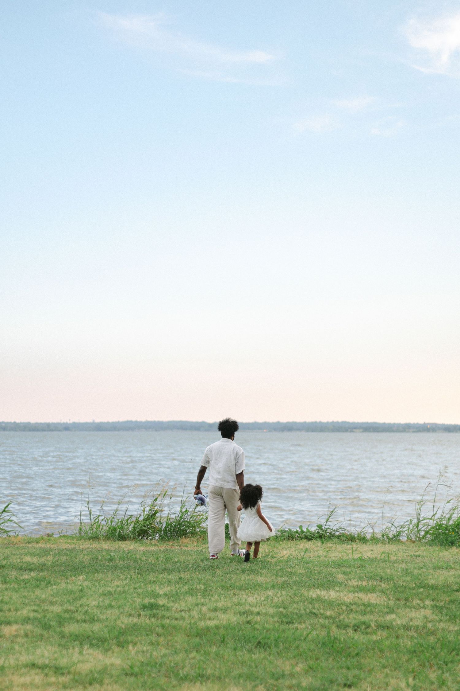 Two people stand on a grassy shoreline overlooking a calm body of water on a sunny day.