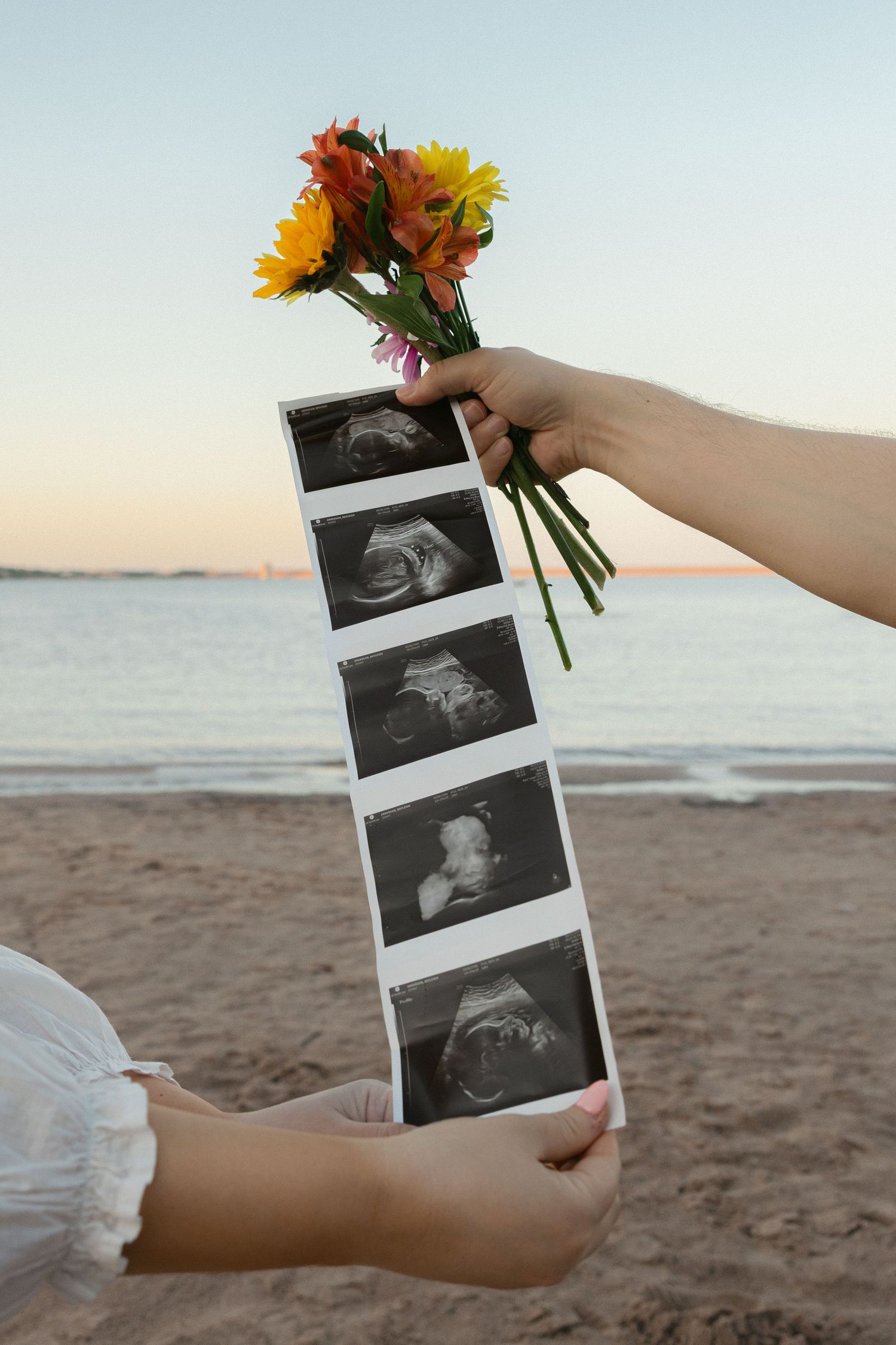 Hands holding ultrasound photos and colorful flowers on a beach at sunset, announcing a pregnancy.
