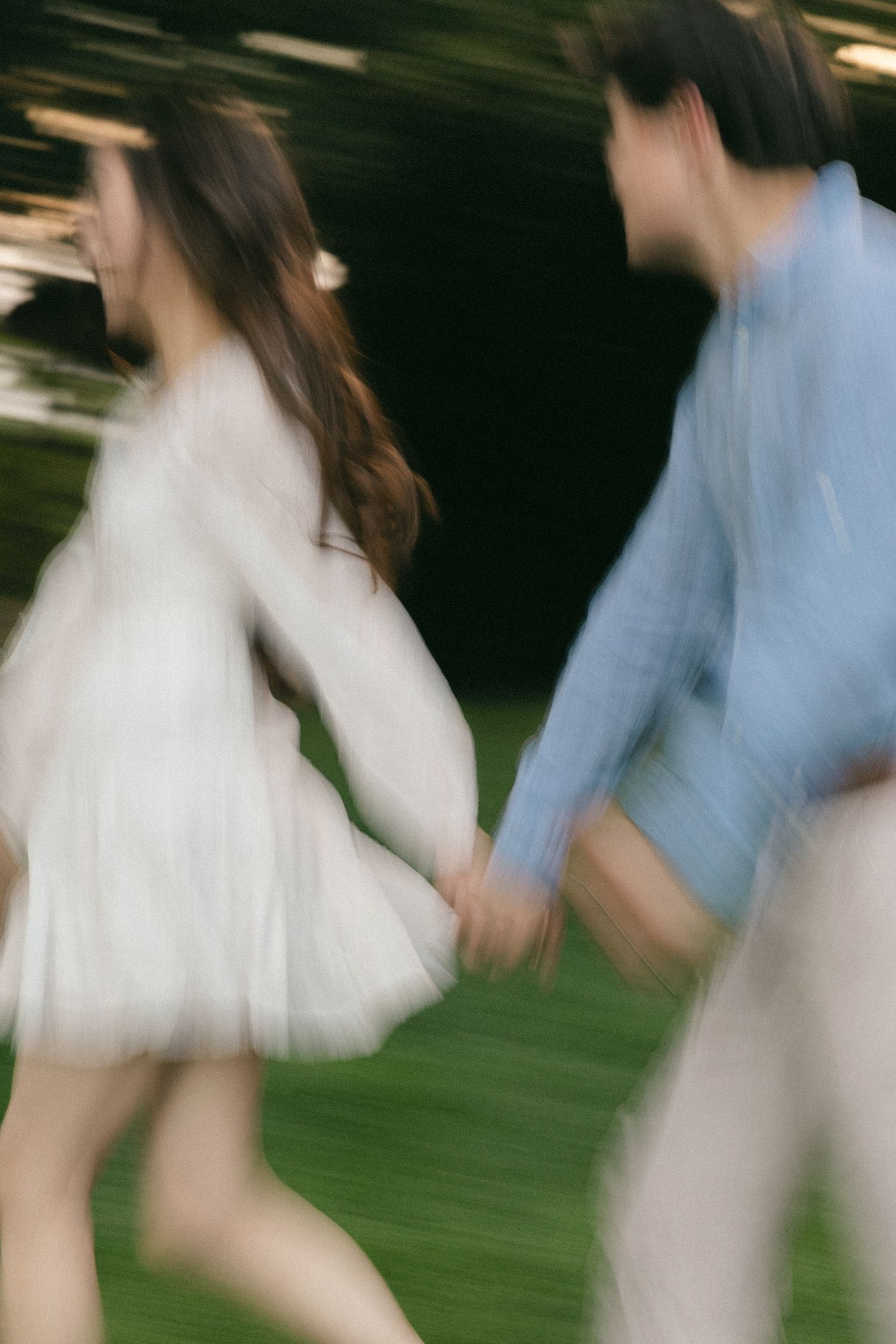 A couple holds hands while walking on grass at night, captured with motion blur in matching white and blue outfits.