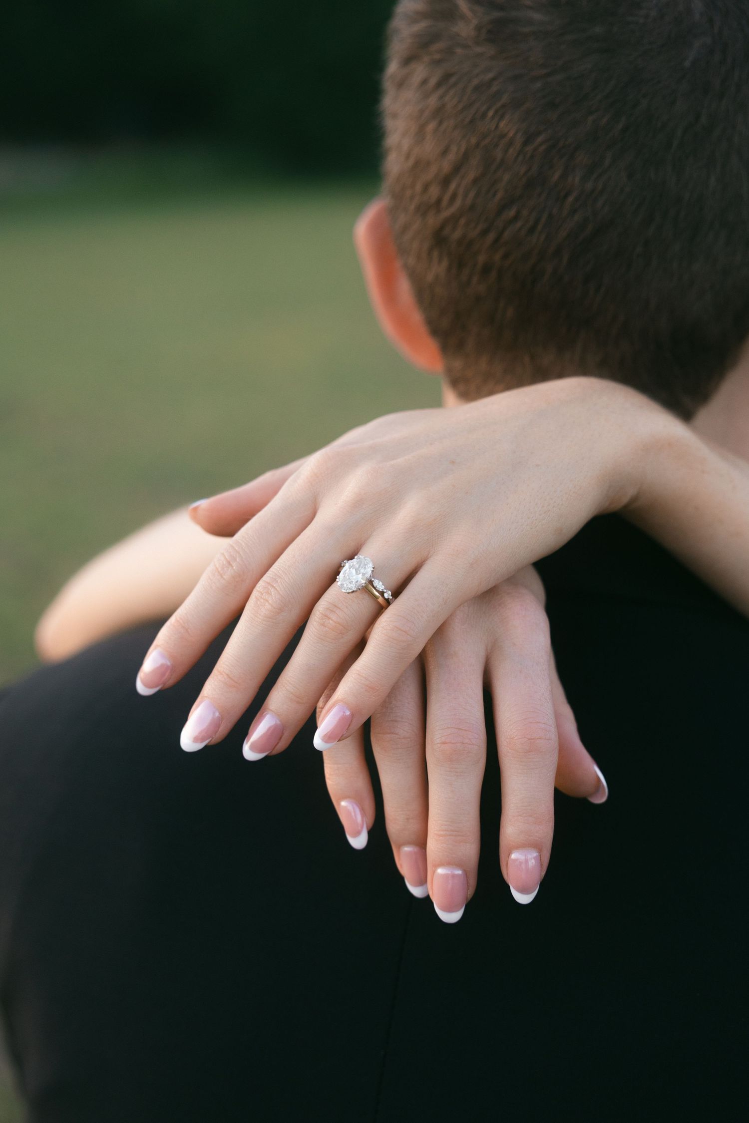 Close-up of bride's hand showing diamond engagement ring resting on groom's shoulder.