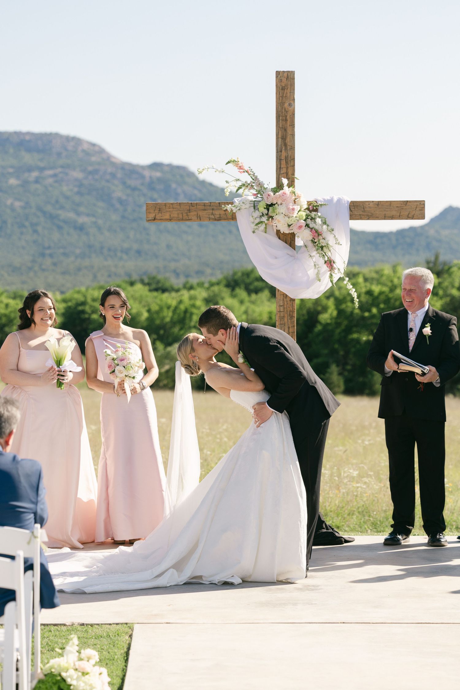 Bride and groom share first kiss at outdoor wedding altar with cross, bridesmaids, and mountain backdrop.