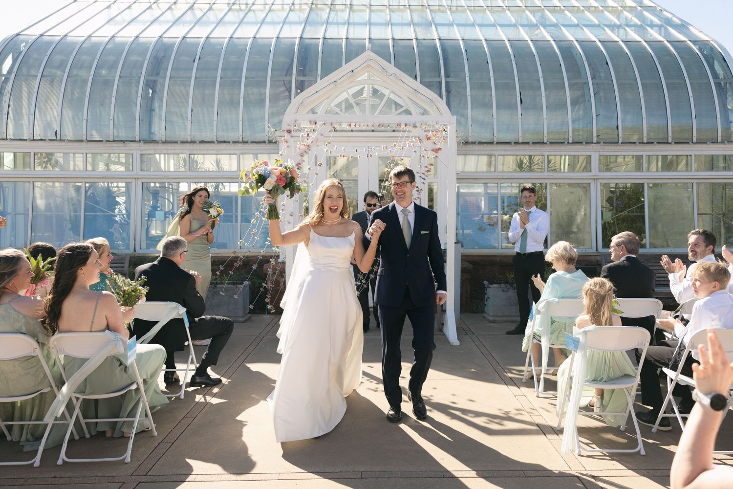 Newlyweds walk down the aisle at a sunlit greenhouse wedding venue as guests celebrate.