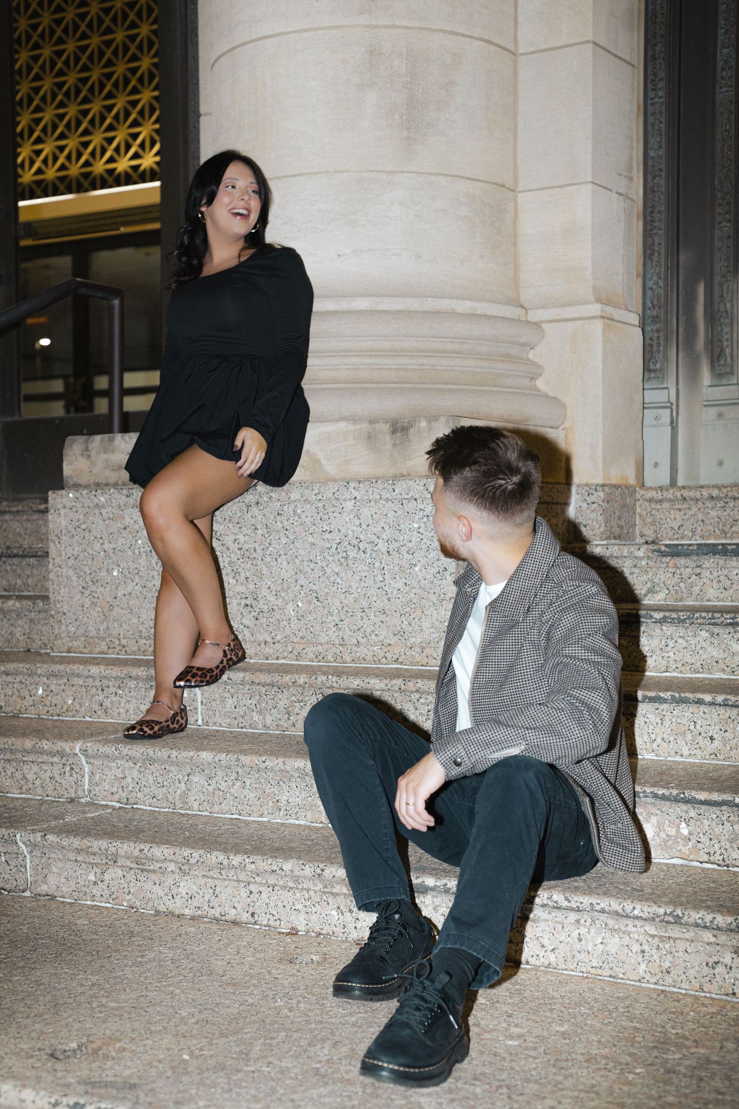 Playful couple posing on stone steps near architectural columns at night.