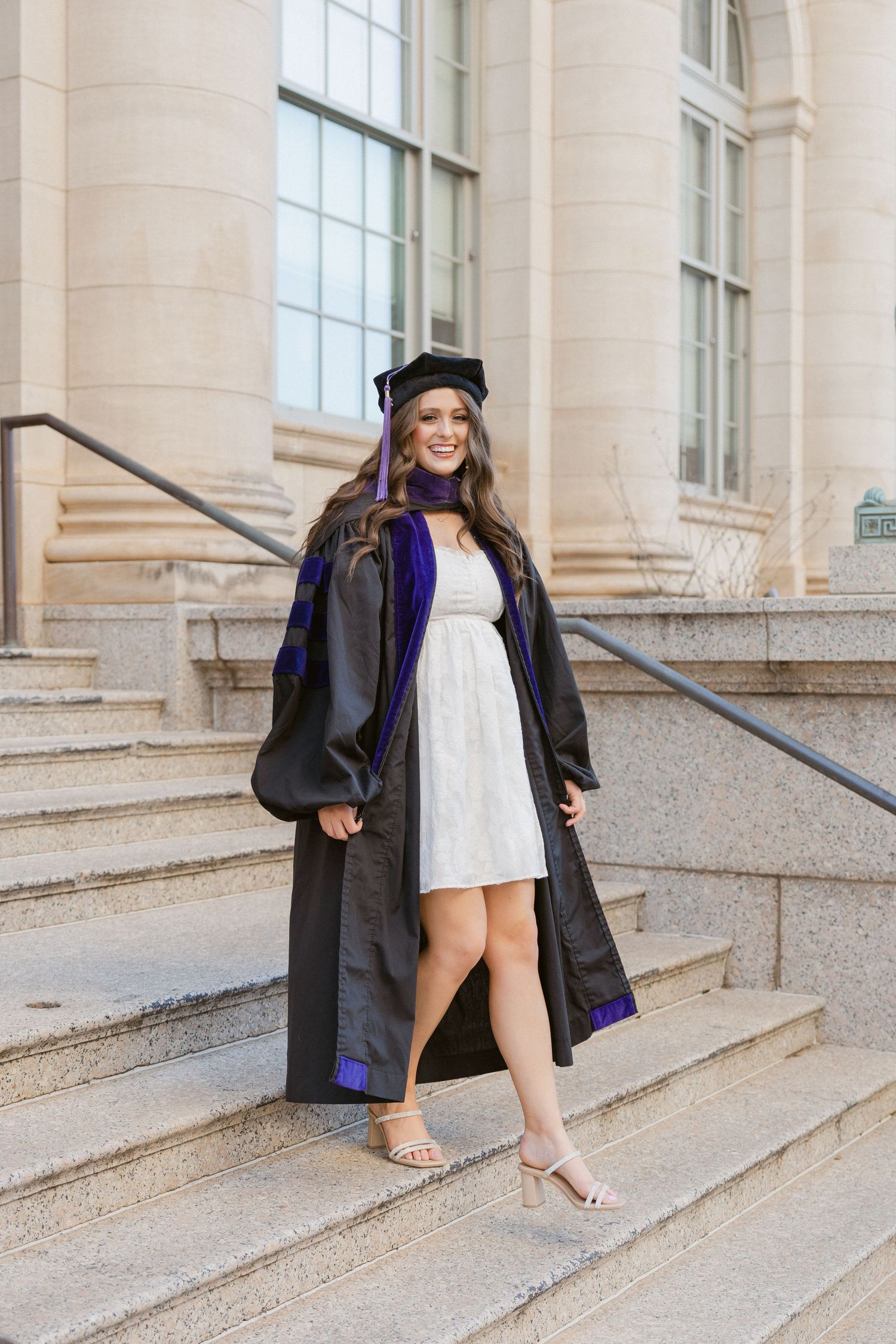 Graduate in cap and gown poses on stone steps in front of a building during graduation.