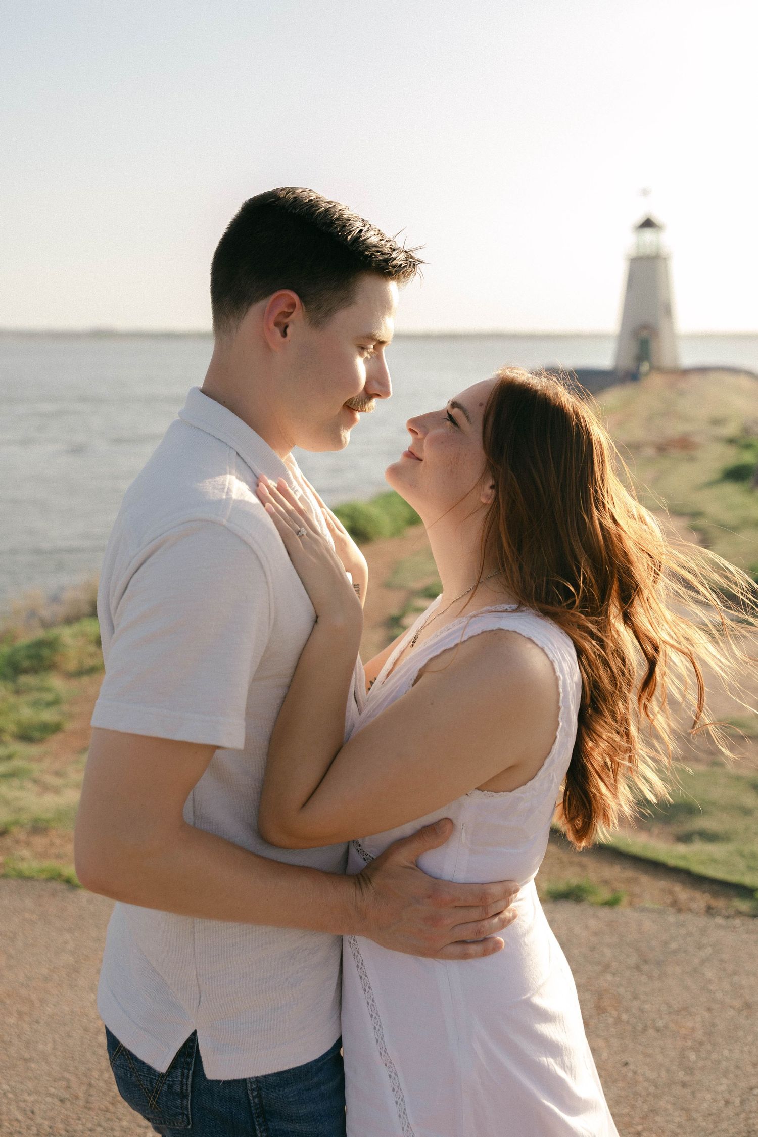 Romantic couple embracing near a lighthouse by the water, gazing into each other's eyes on a sunny day.
