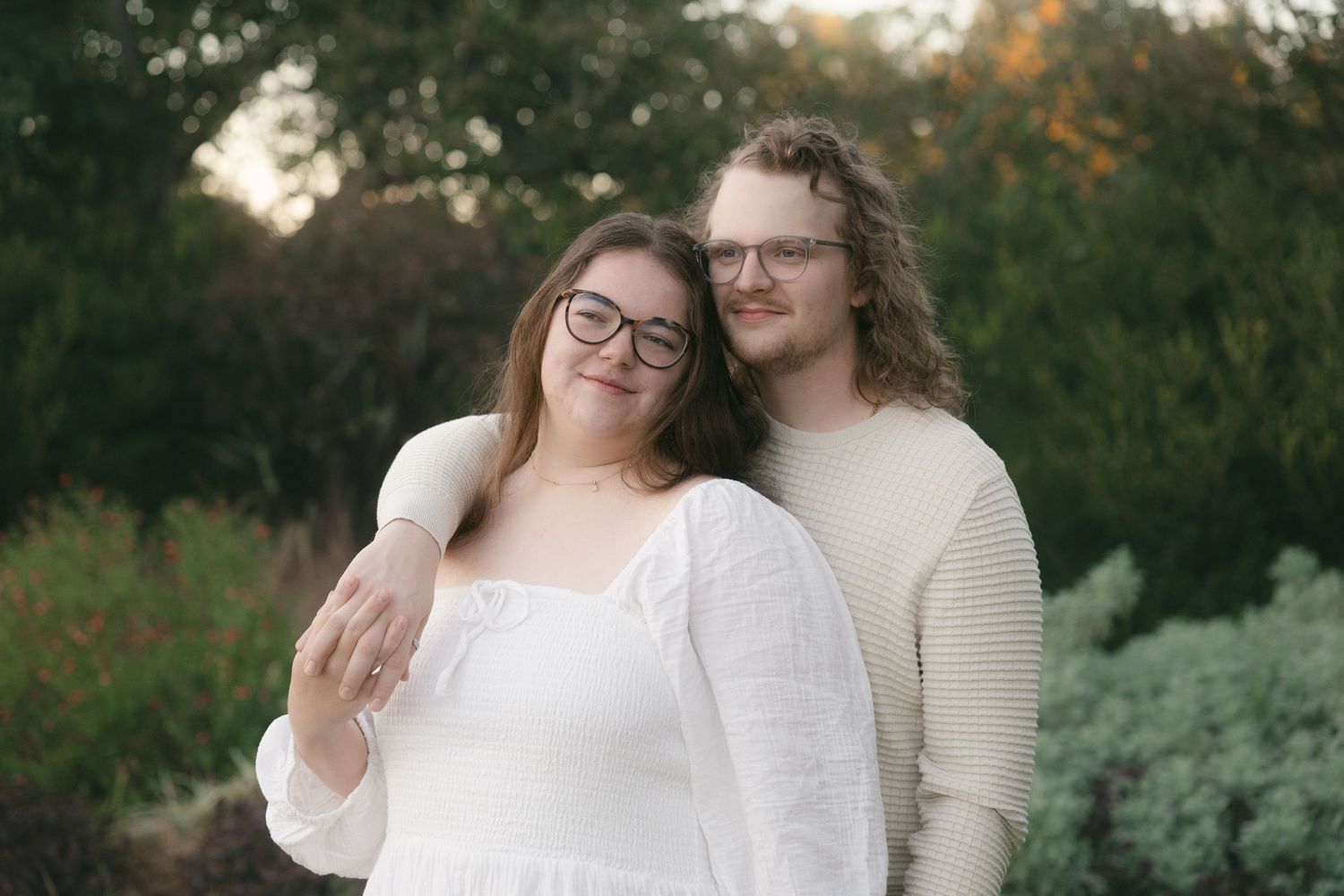 A couple in white outfits embraces during an outdoor autumn photoshoot with natural greenery in the background.