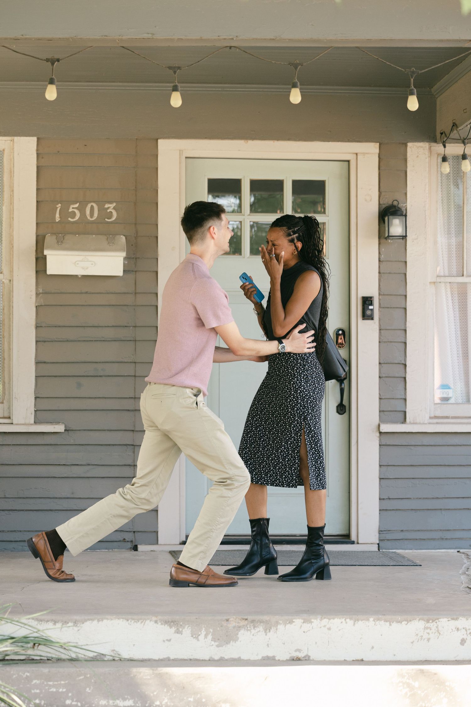 Man proposing to woman on house porch numbered 1503, she covers her mouth in surprise holding a small blue box.