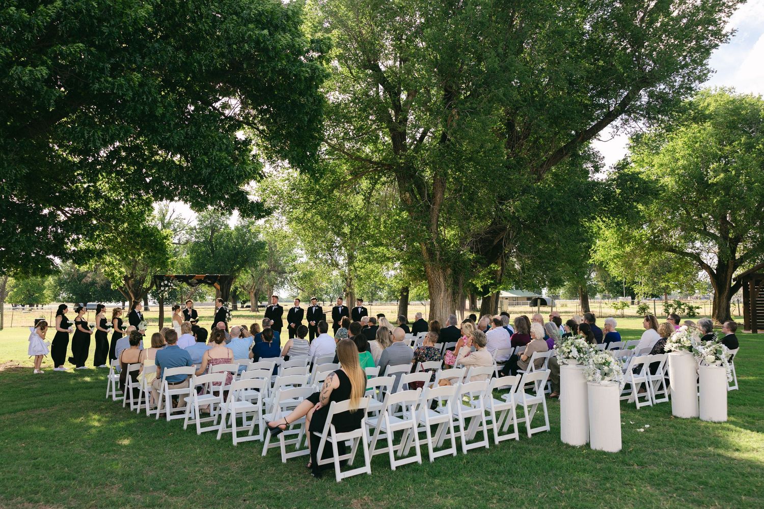Outdoor wedding ceremony with guests seated in white chairs on lush green lawn under large shade trees.