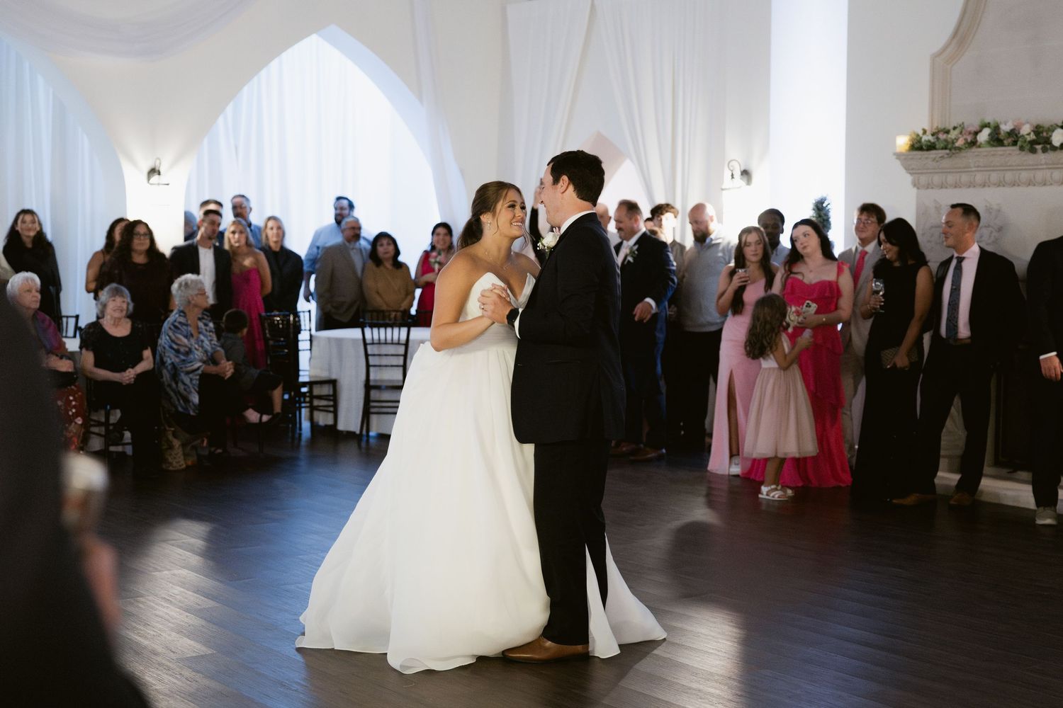 A couple shares their first dance surrounded by guests in an elegant white-draped venue.
