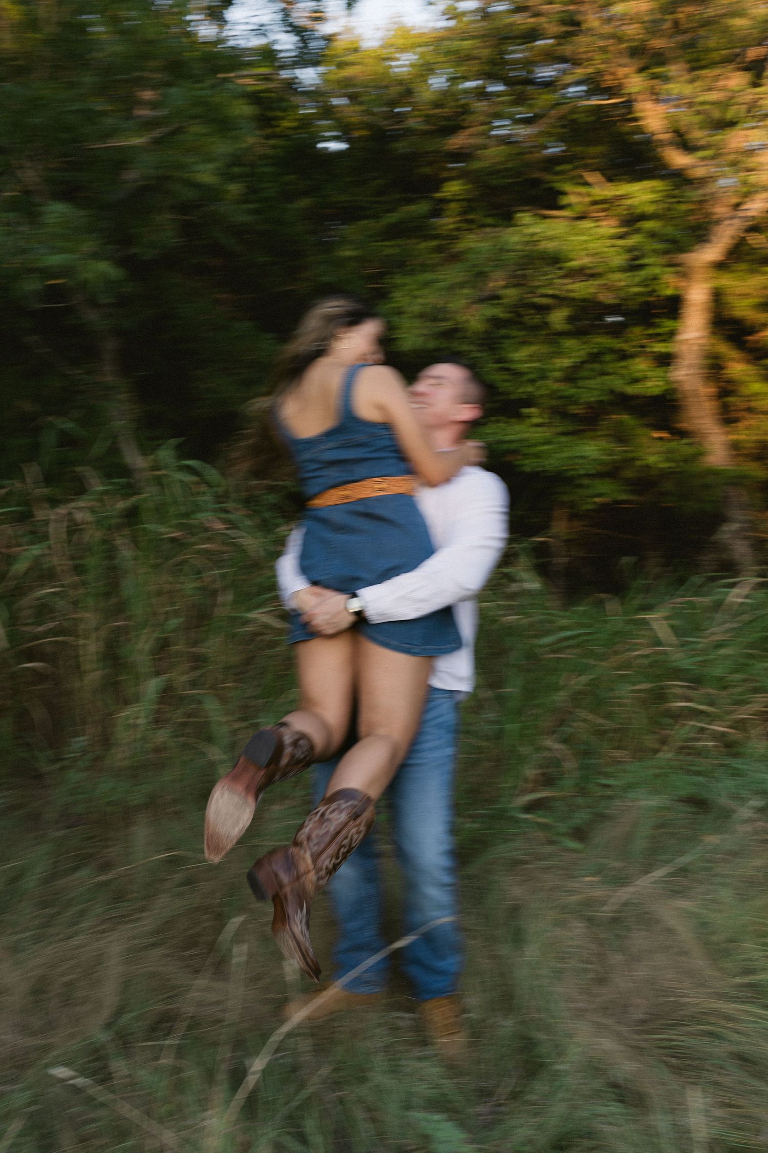 A couple embraces playfully in a field with tall grass and trees during golden hour.