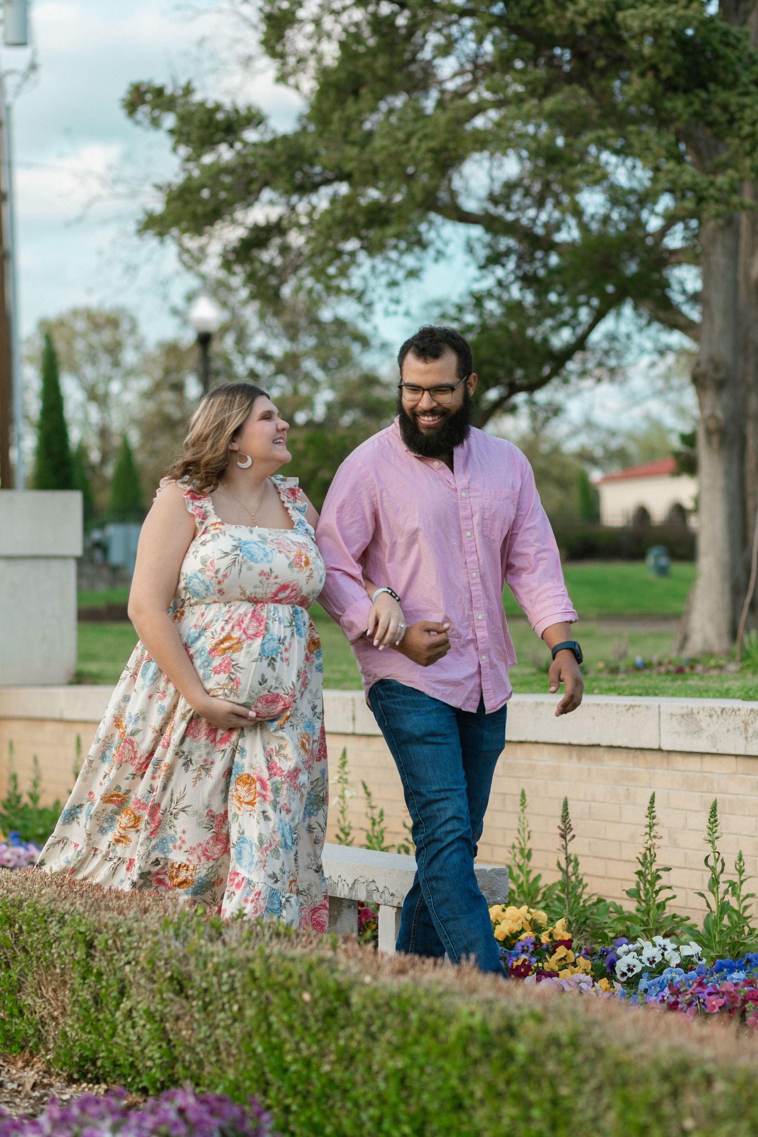 A happy couple strolling through a blooming garden with colorful flowers, she in a floral dress and he in a pink shirt.