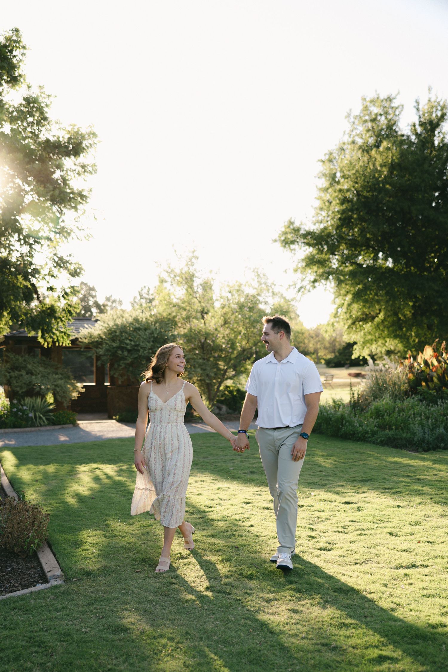 A couple holds hands walking through a sunny green garden, woman in floral dress, man in white polo and khakis.