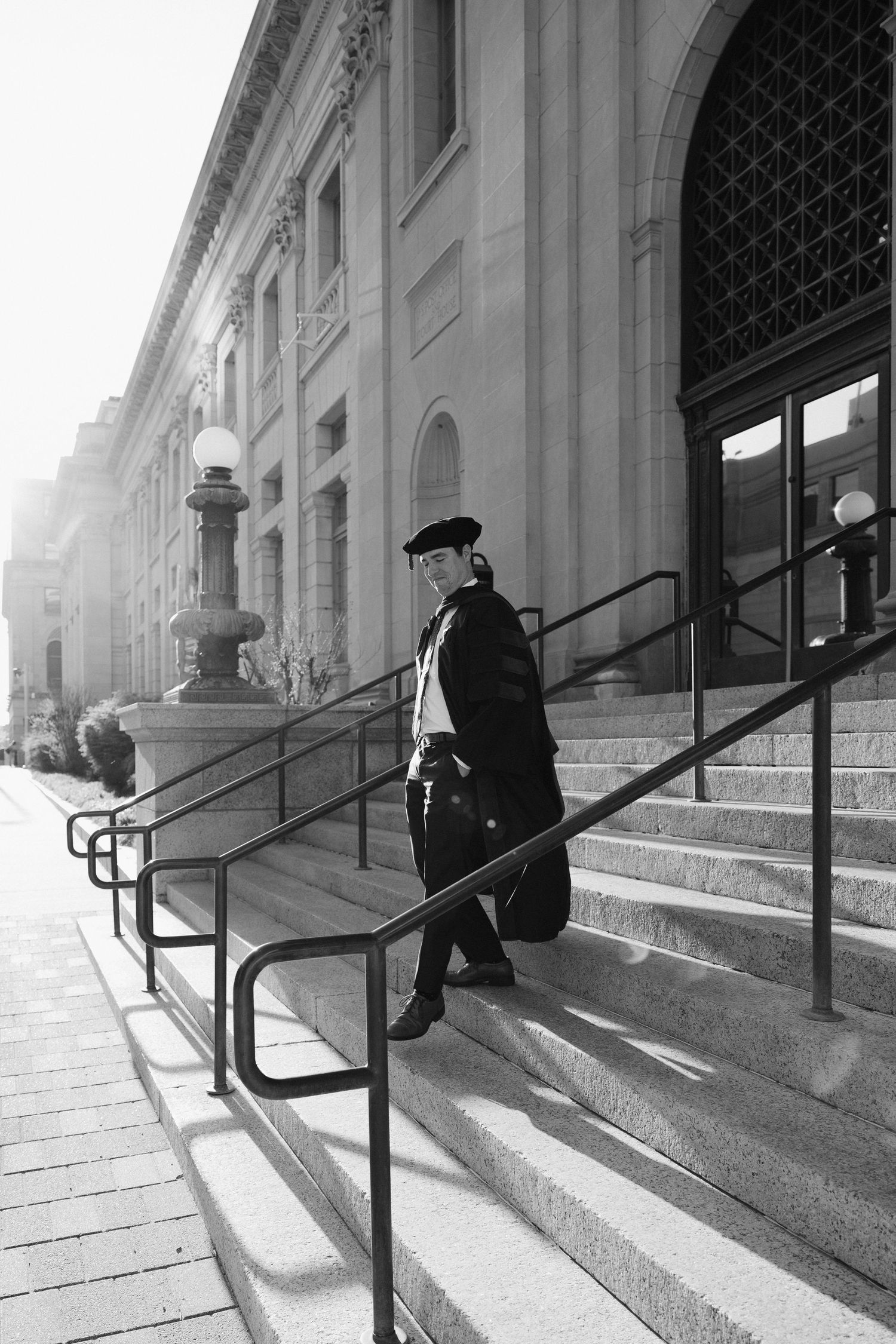 Graduate in academic regalia descending stone steps of a classical building in black and white.