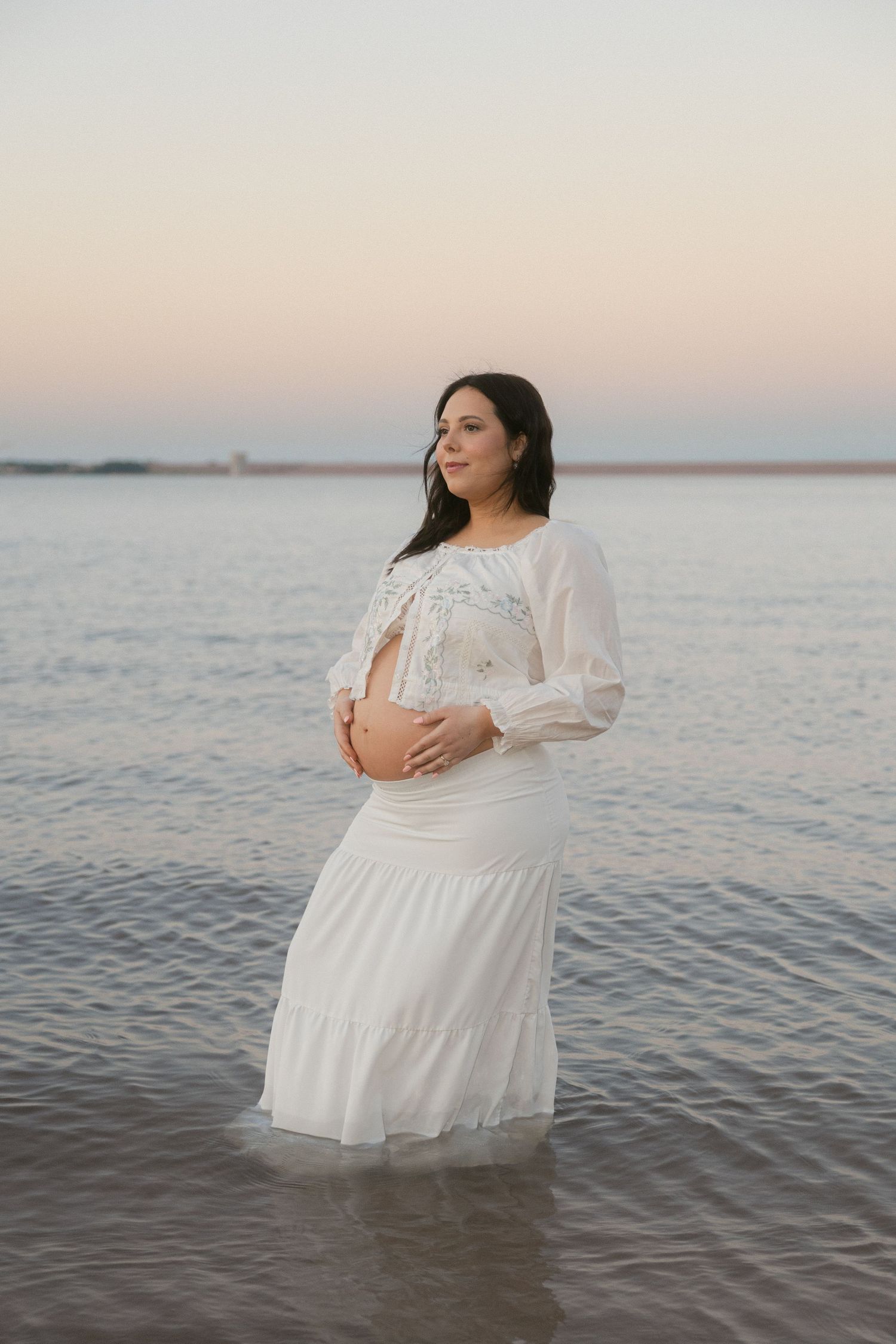 Pregnant woman in white dress standing in shallow ocean water at sunset, cradling her baby bump.