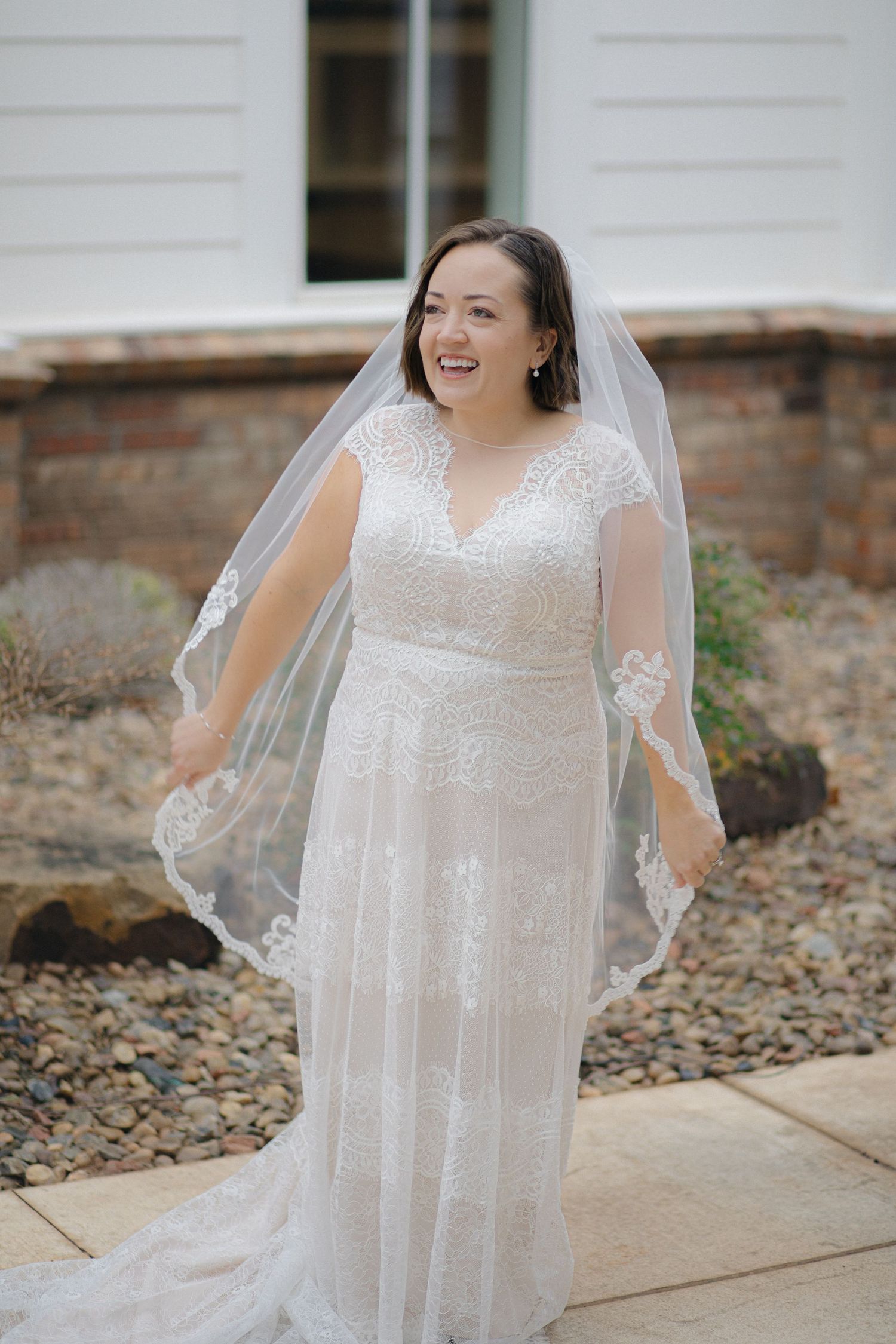 A bride in a flowing white lace wedding dress and veil stands outdoors against a brick wall background.