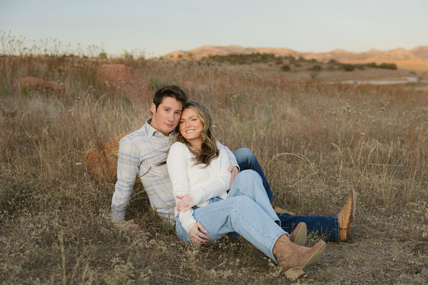 A couple sits together in a golden field during sunset, sharing intimate moments across multiple poses.