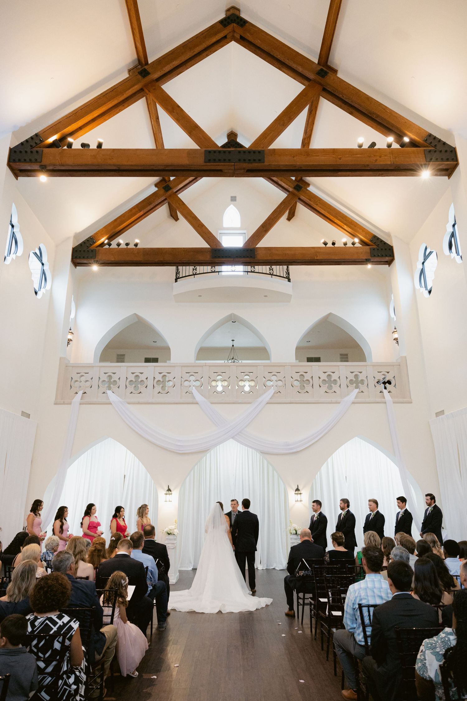 Indoor wedding ceremony in a bright white chapel with exposed wooden beams and gothic arched windows.