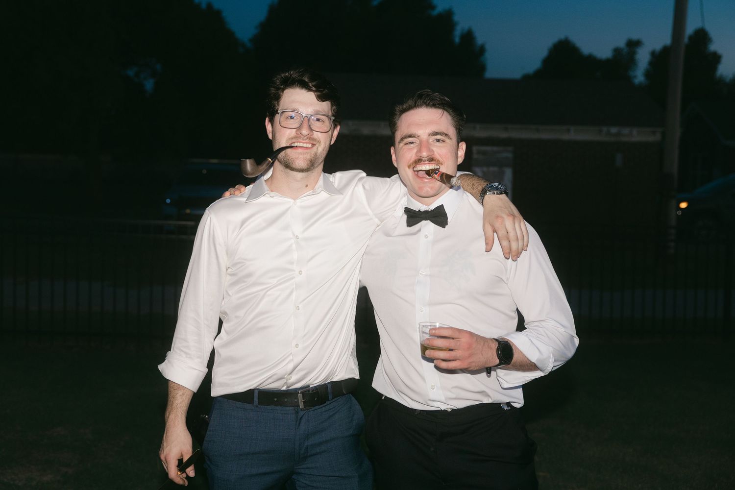 Two smiling men in white dress shirts pose together outdoors at dusk, one wearing a bow tie.