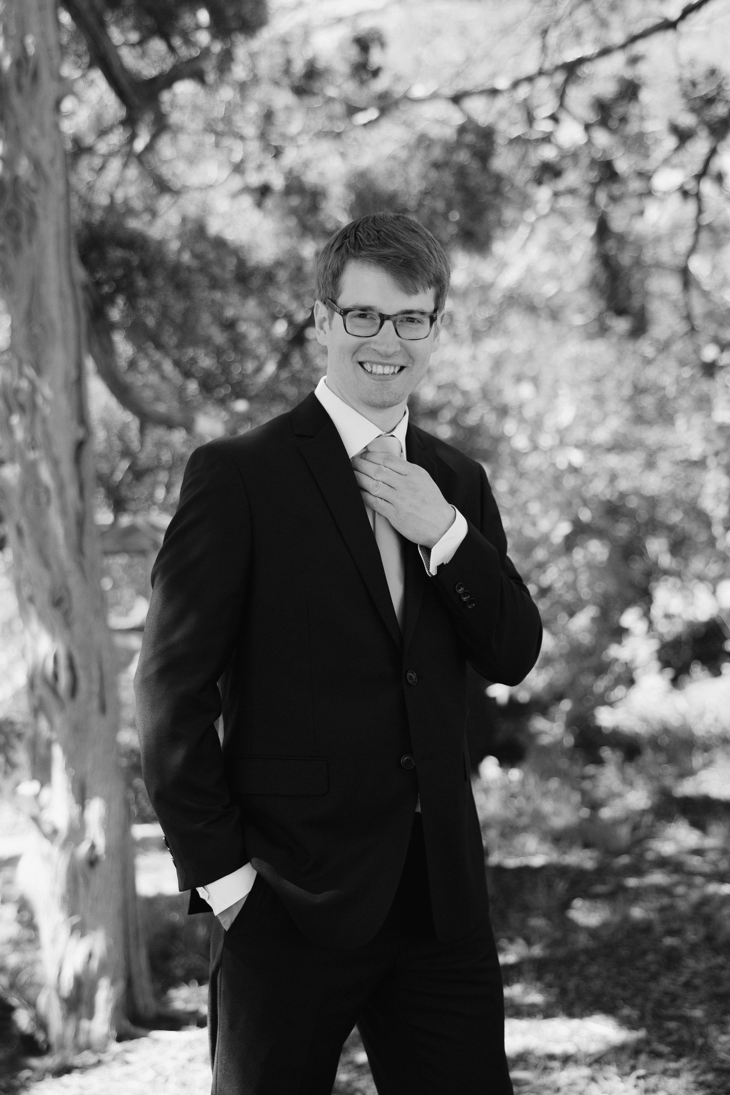 Smiling groom in black suit and glasses adjusting his tie outdoors in a black and white portrait.