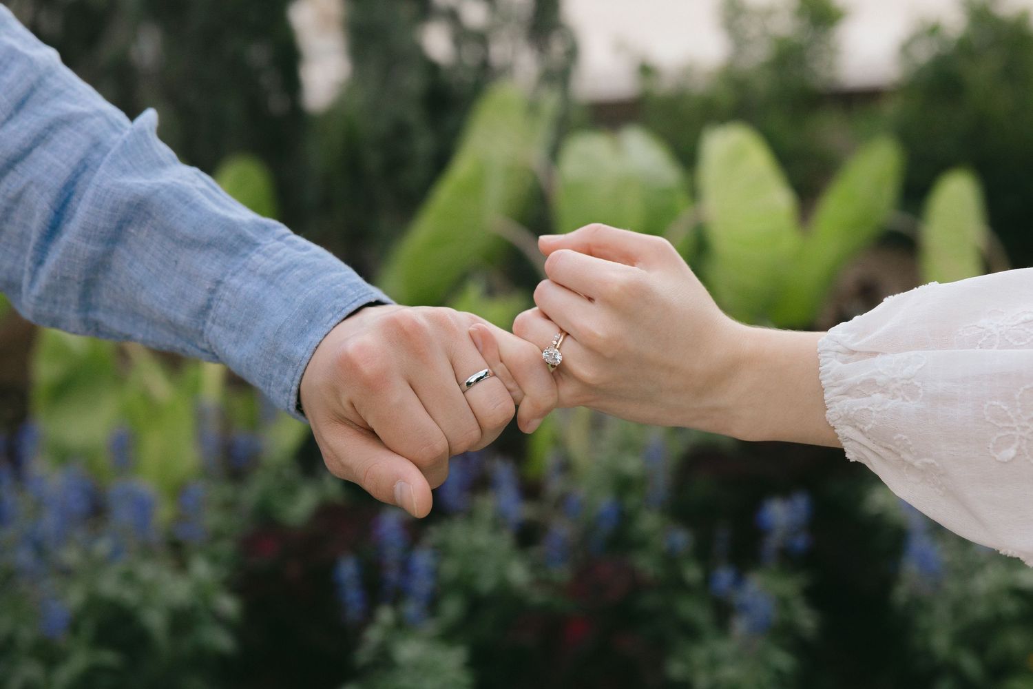 Close-up of an engaged couple holding hands outdoors, showcasing a diamond engagement ring among garden flowers.