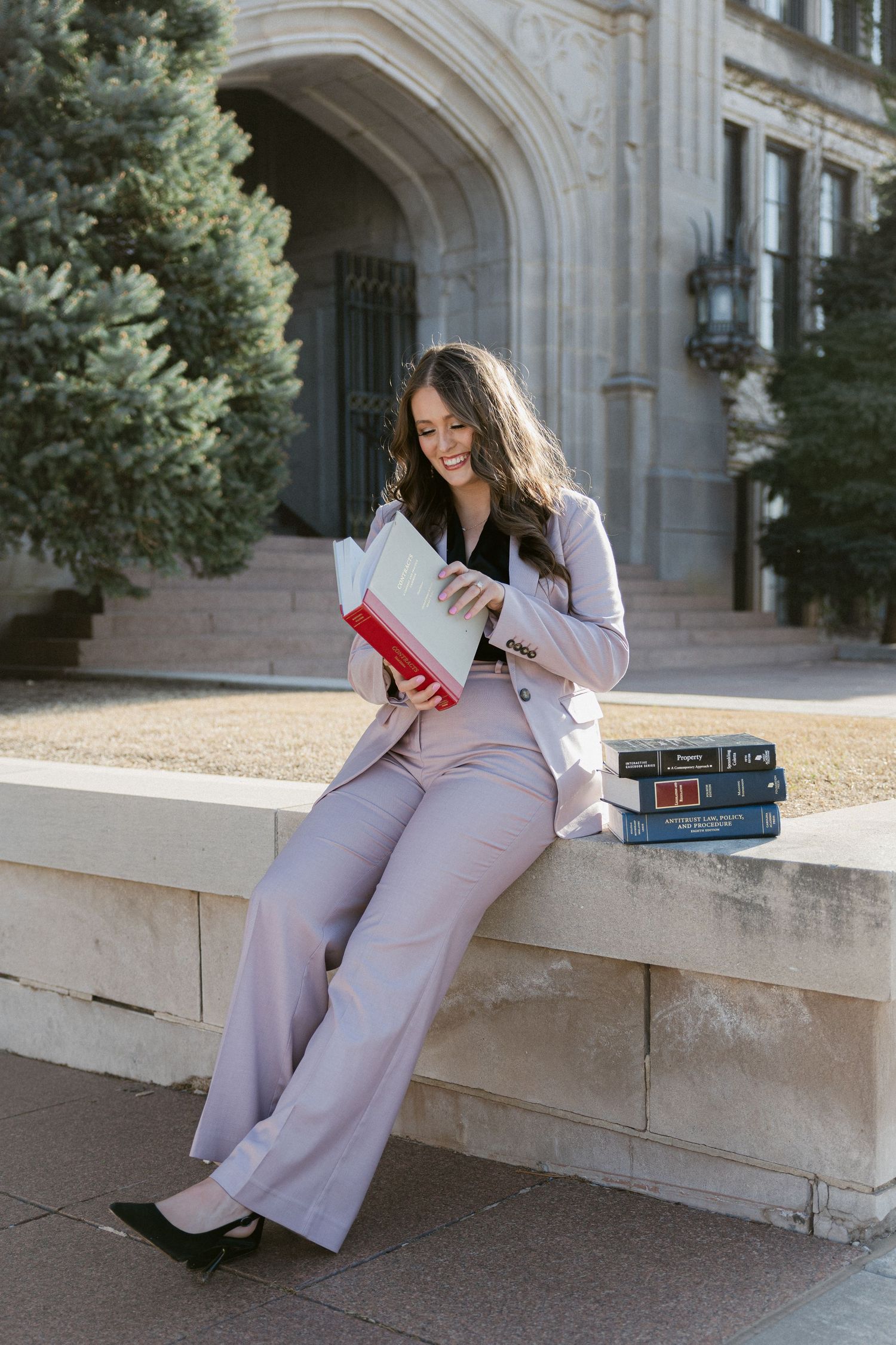 Someone in a light-colored pantsuit sits on a stone wall while reading a red book in an elegant outdoor setting.