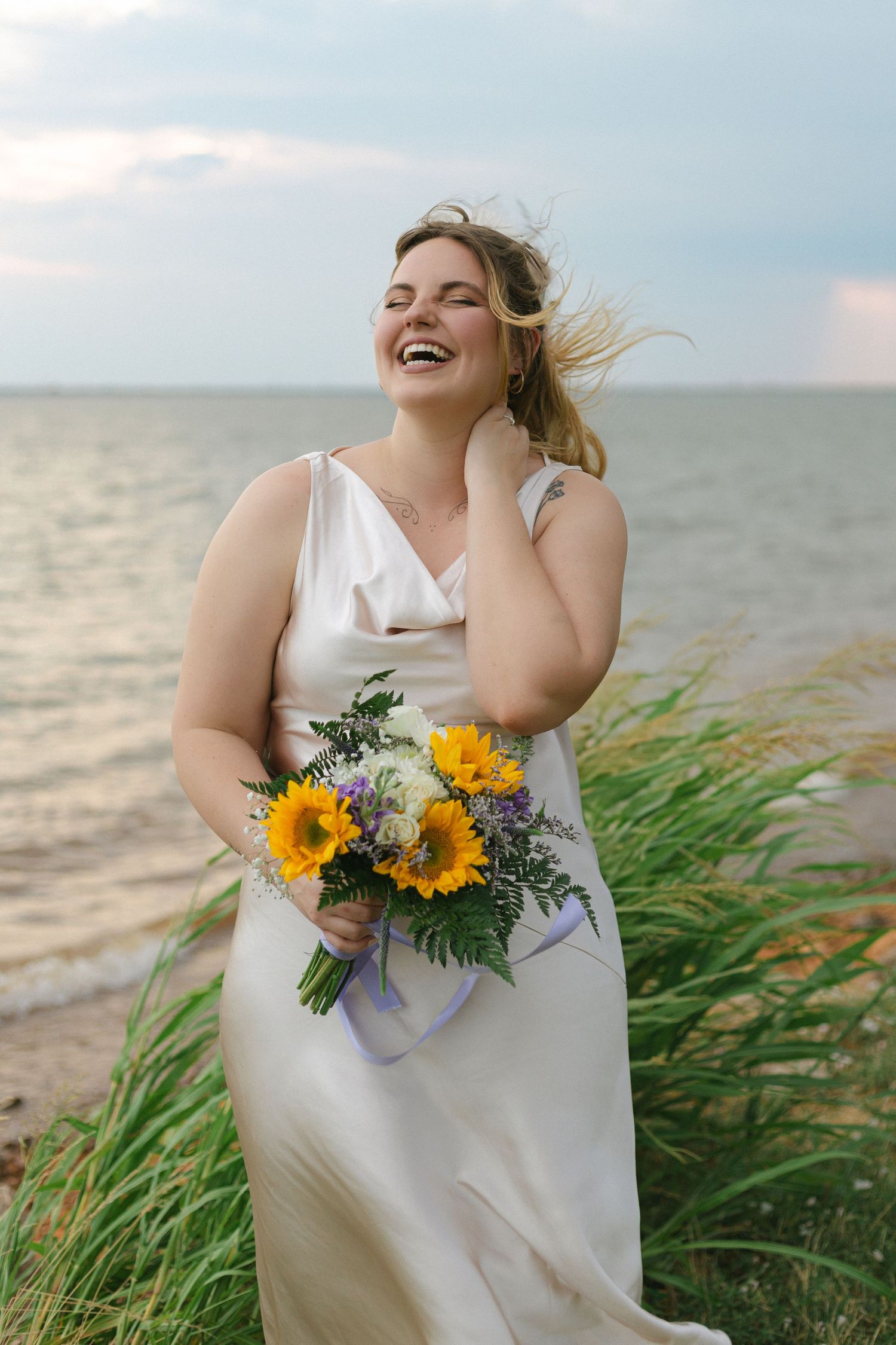 Woman in flowing white dress stands on beach with sunflower bouquet as wind blows through tall grass at sunset.