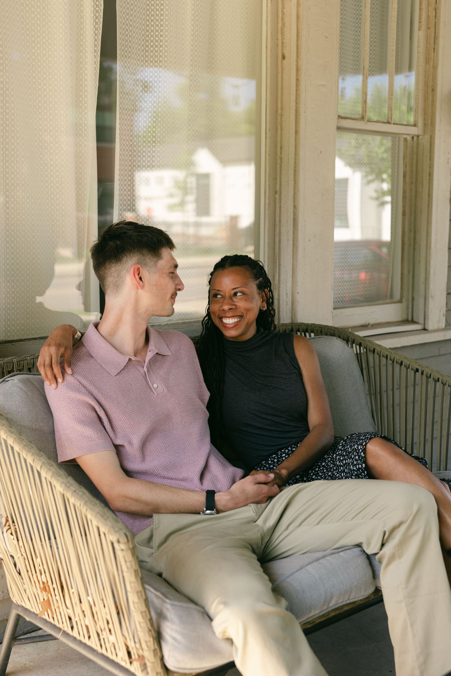 A smiling interracial couple sits together on a wicker porch sofa, the man in pink polo embracing the woman in black top.