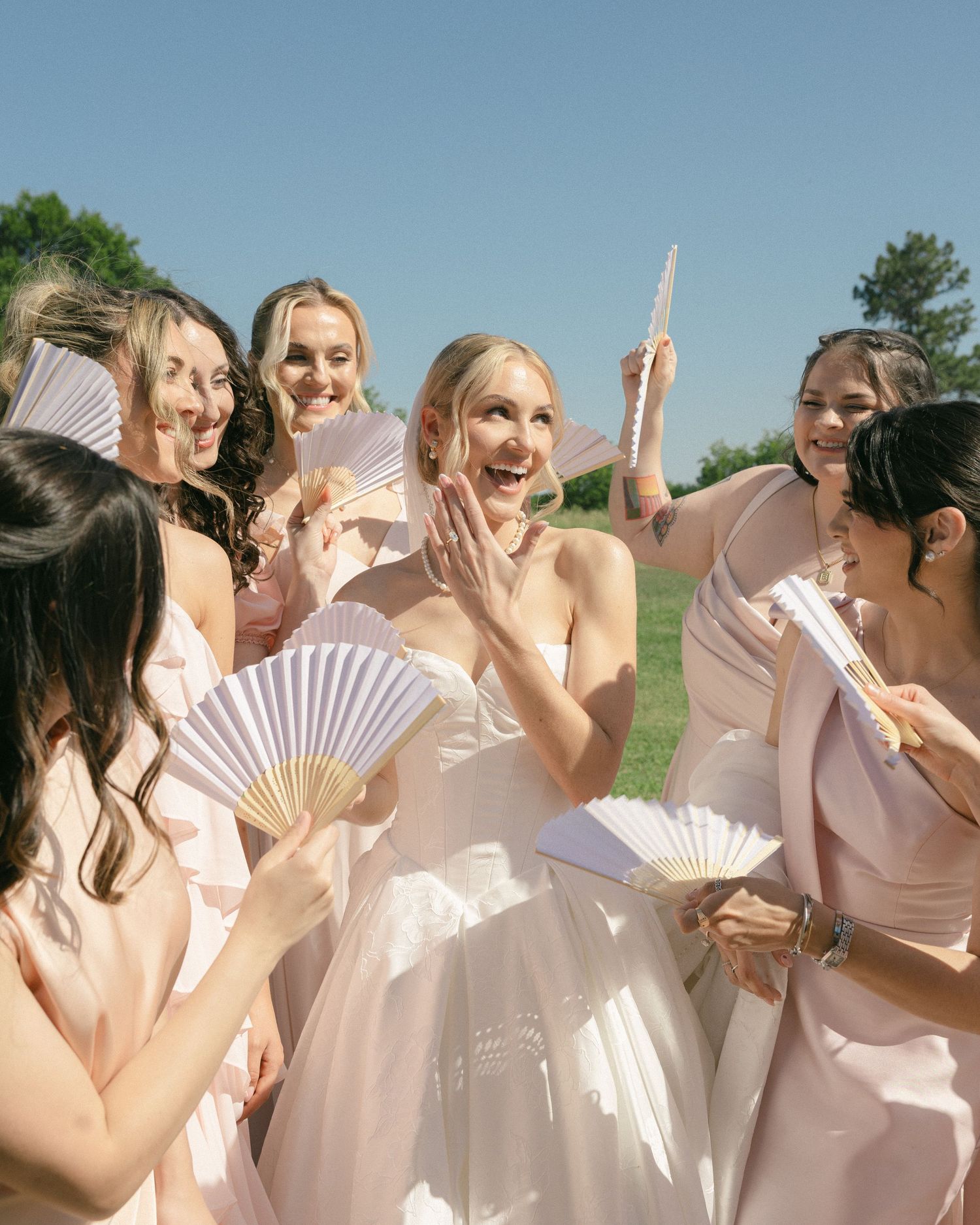 Excited bride in white gown laughing with bridesmaids in blush dresses holding paper fans outdoors.