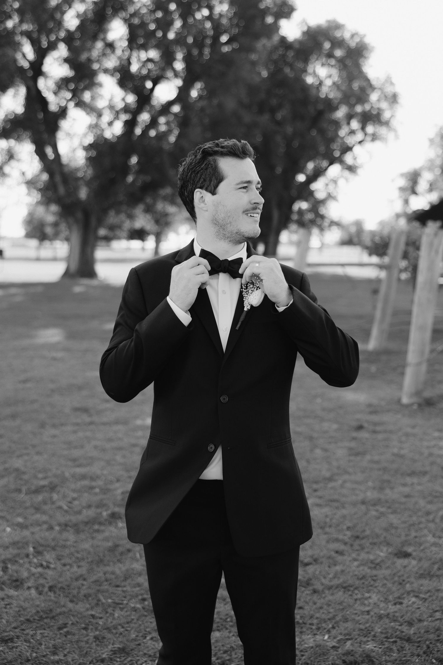 Dapper groom in black tuxedo and bow tie smiling outdoors in a classic black and white wedding portrait.