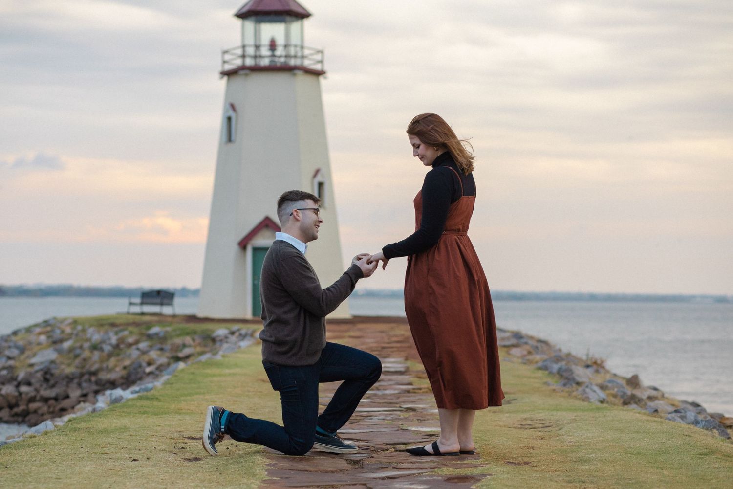 A romantic engagement session at a seaside lighthouse during sunset with waves crashing on the rocky shore.
