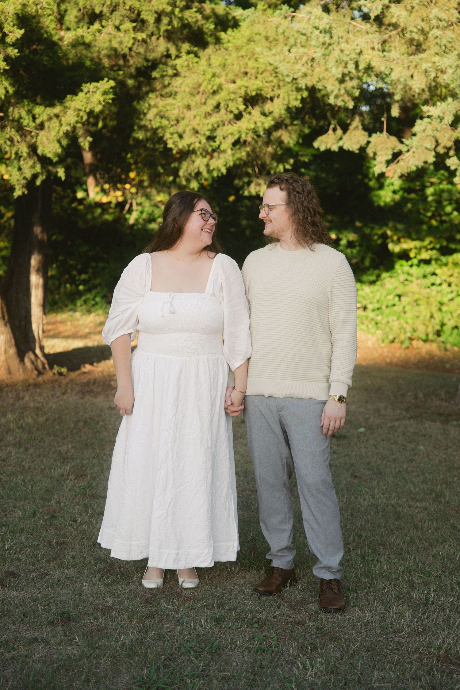Two people in white and gray outfits stand together on grass with green trees in the background.
