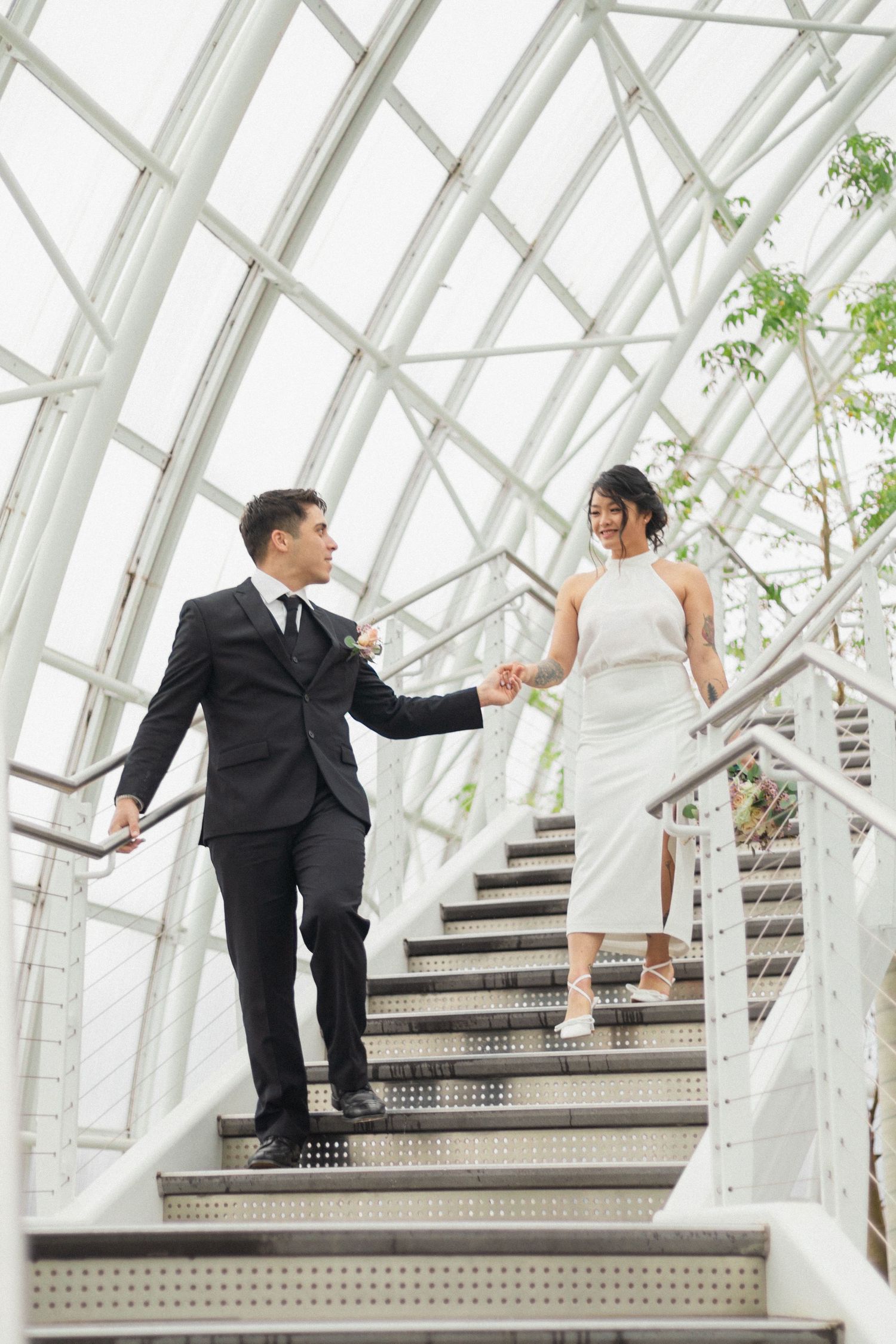 Elegant couple descending white metal stairs in a modern glass-encased structure.