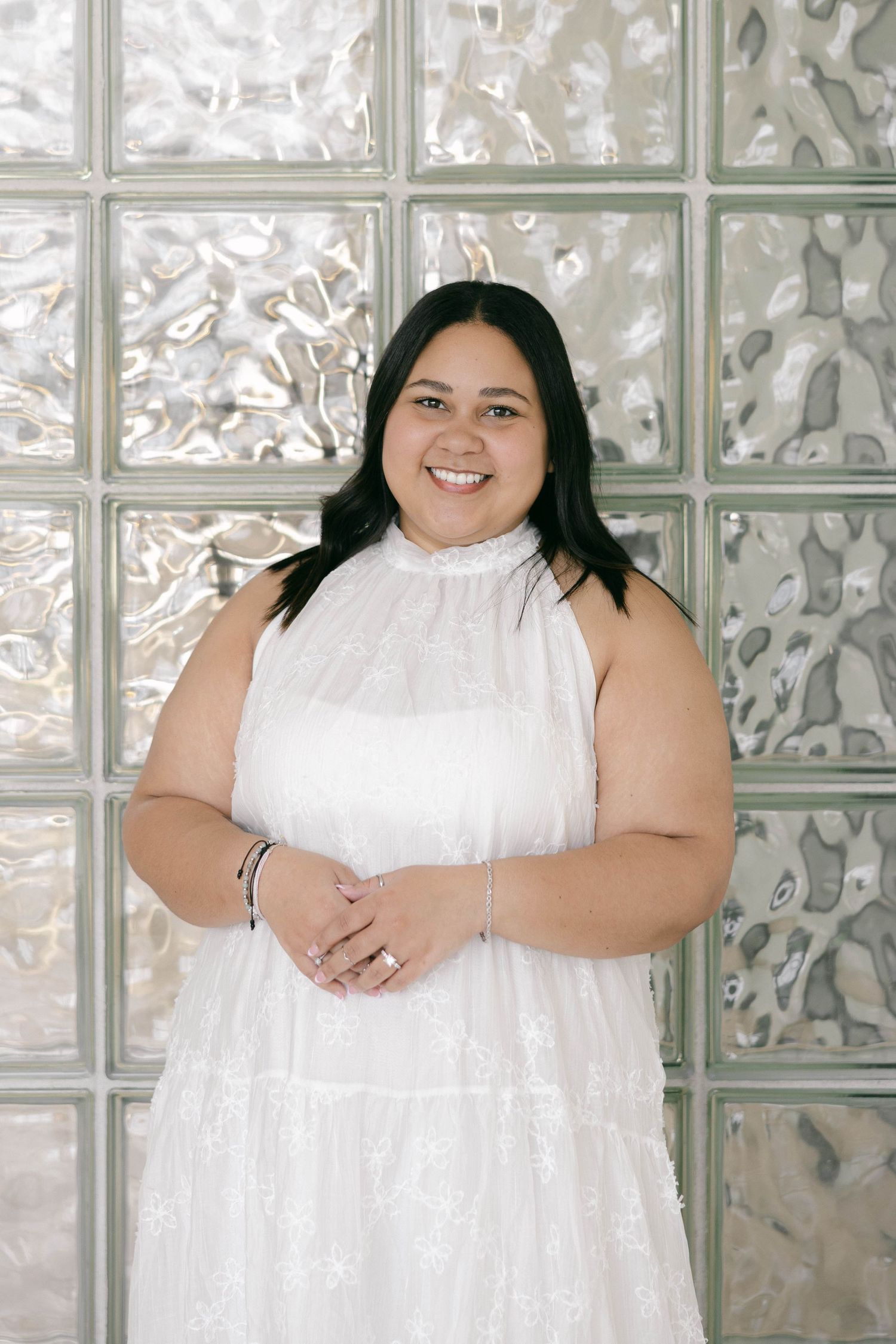 Smiling woman in white sleeveless dress poses in front of glass block wall.