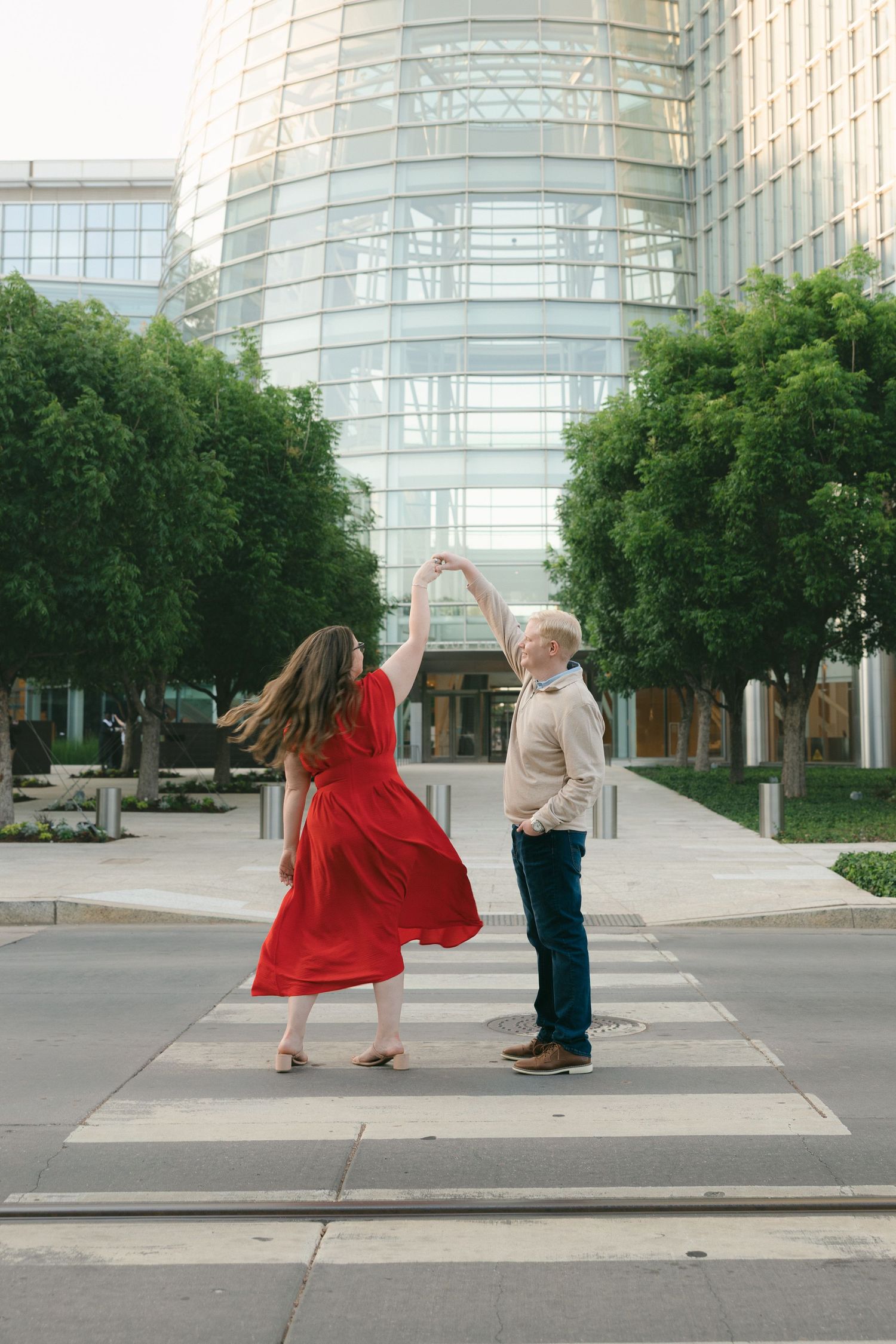 Couple dancing on a crosswalk in red dress and tan suit, with modern glass building and green trees backdrop.