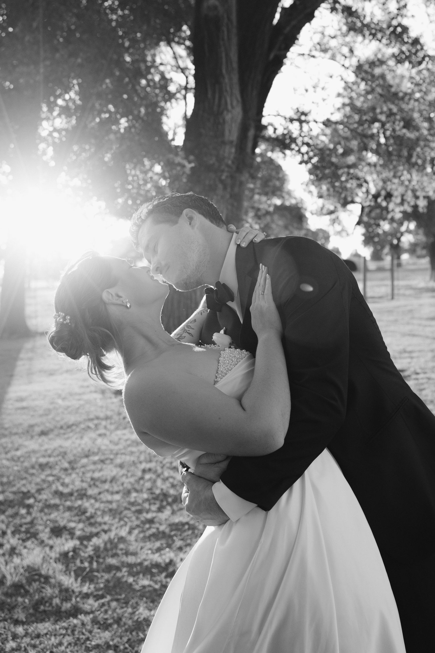 Bride and groom share romantic dip kiss in sunlit park in black and white wedding photo.