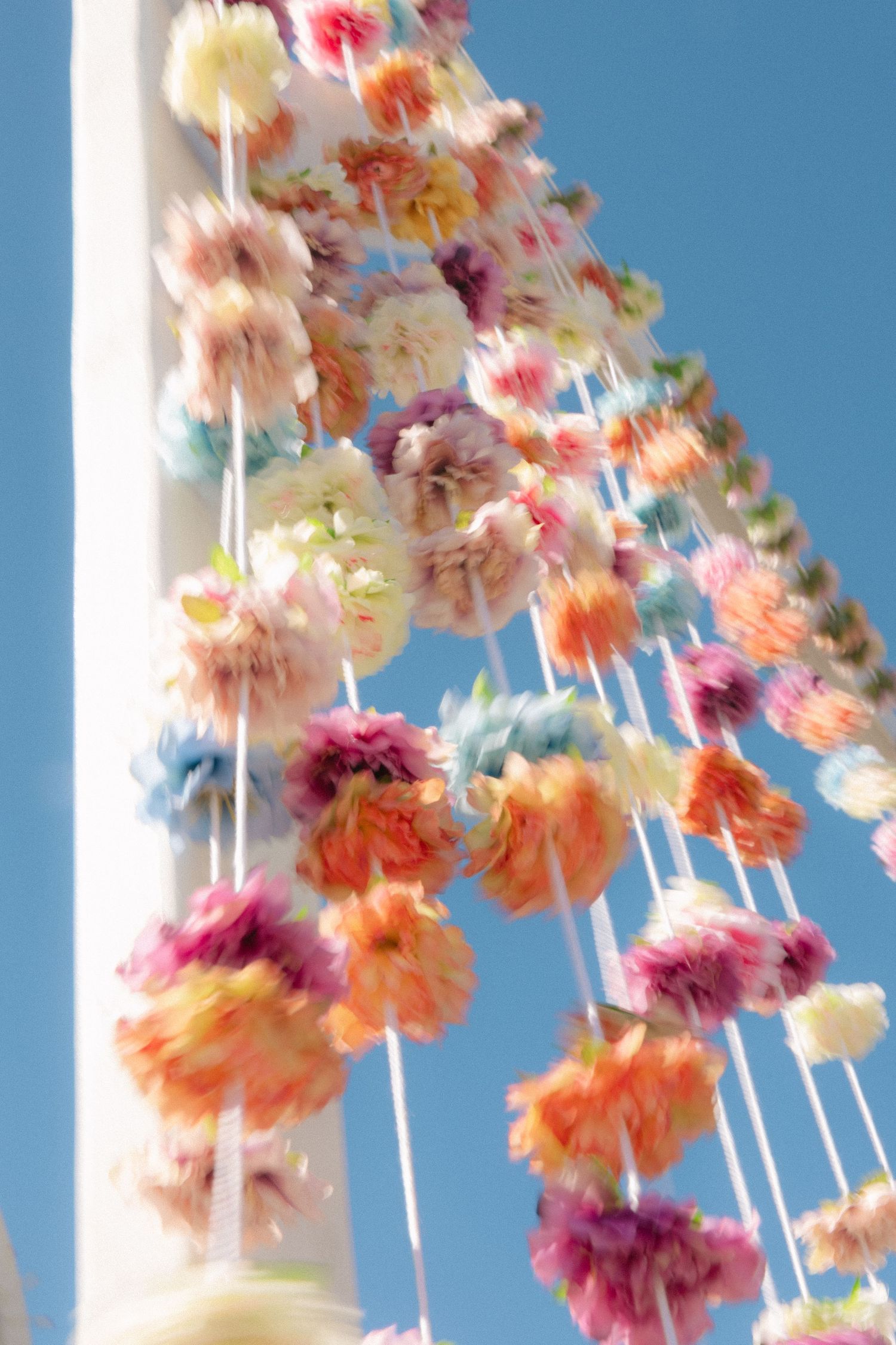 Colorful floral garlands with pink, orange, and cream flowers hanging from a white pole against a blue sky.