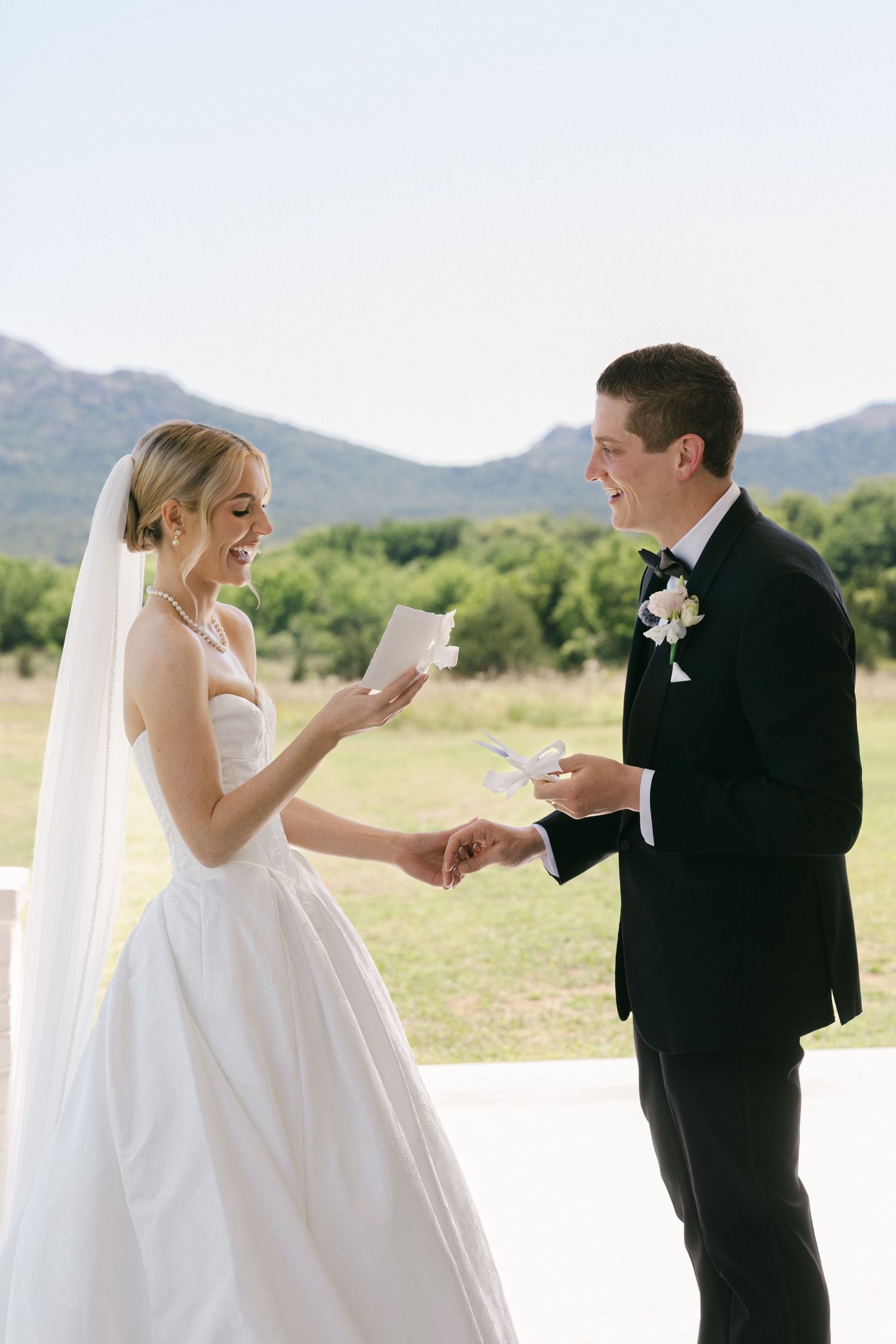 Bride and groom exchange vows outdoors holding cards, smiling, with green hills and blue sky in background.