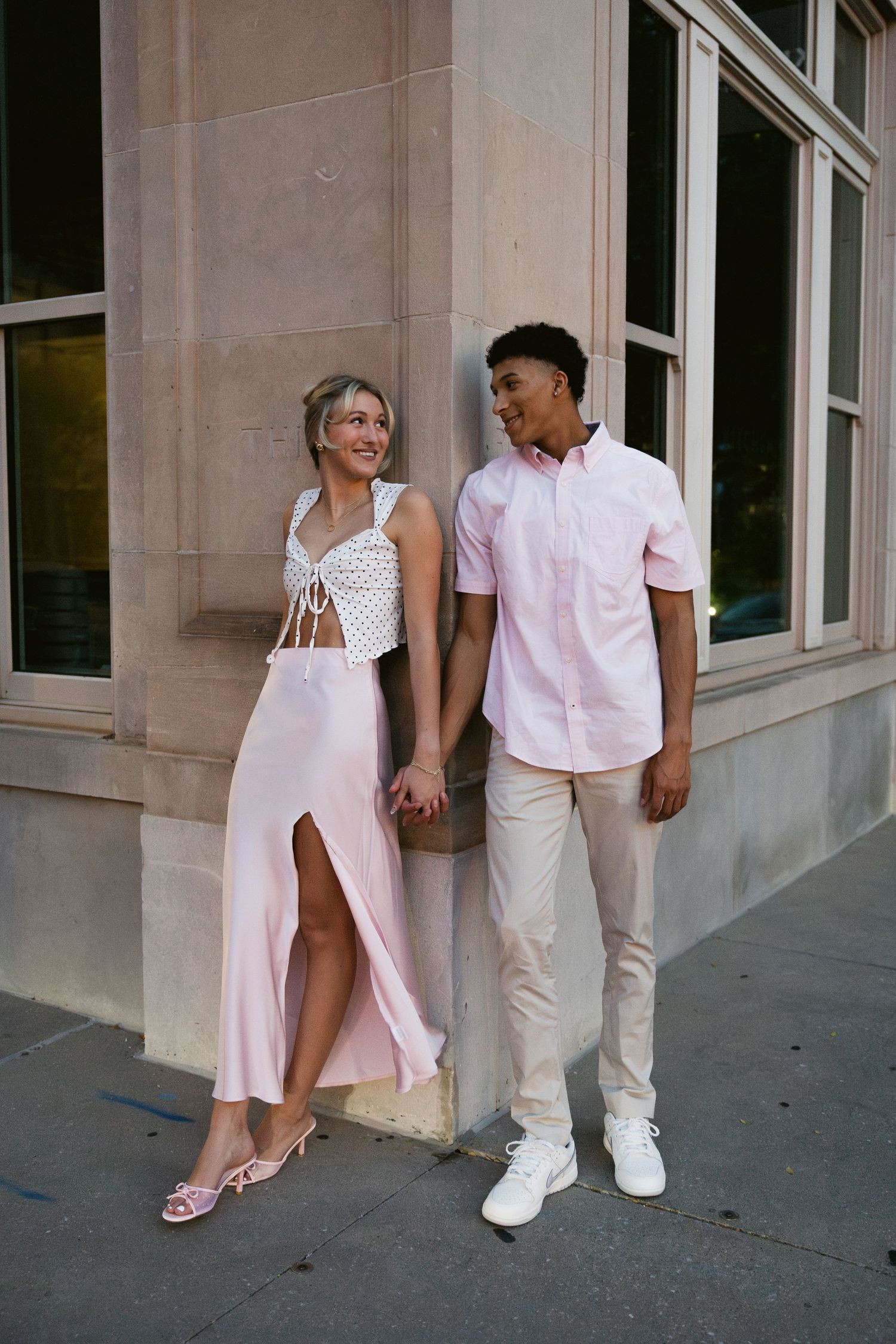 A couple in coordinated pink and white outfits hold hands while standing against a building wall.