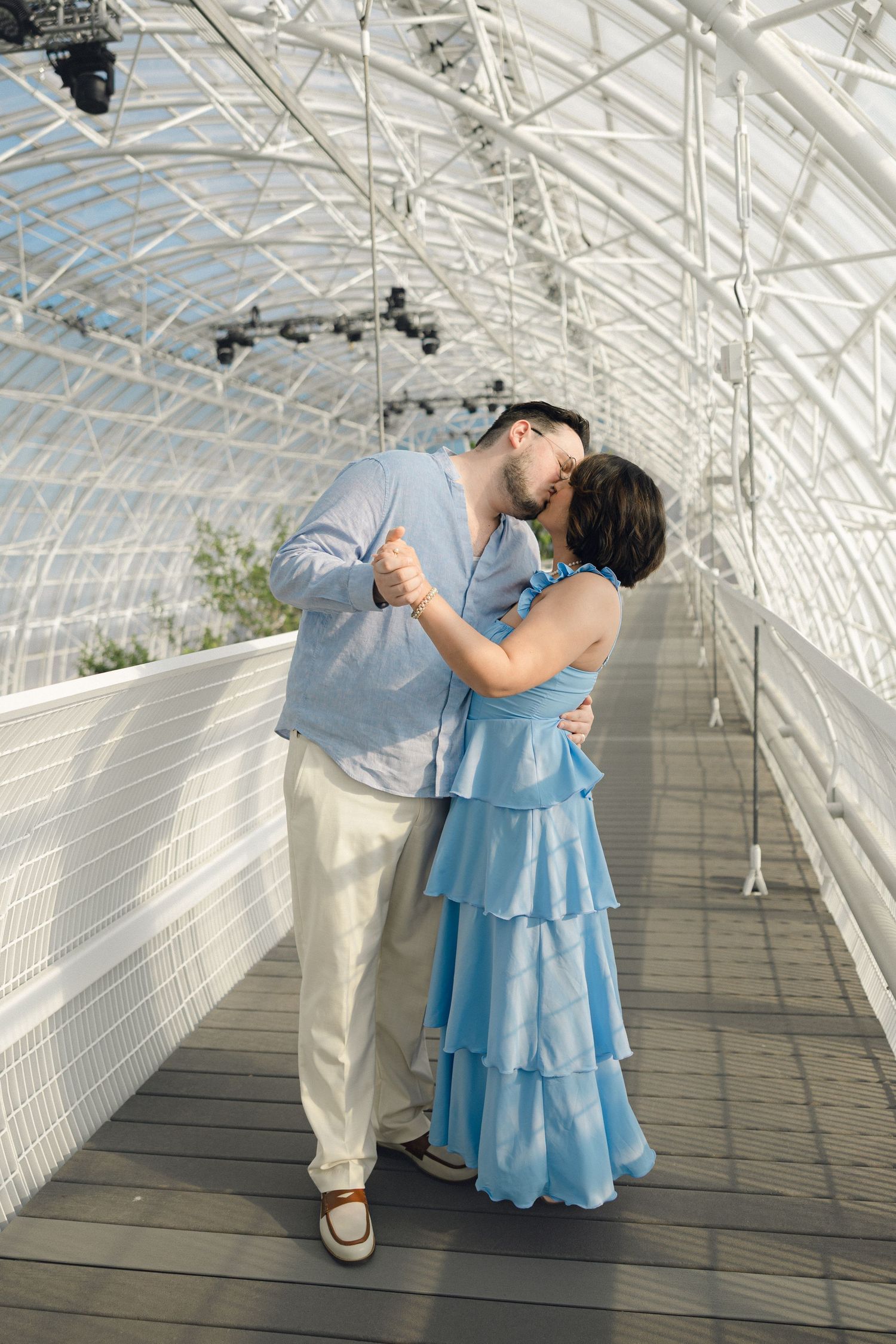 Couple sharing a romantic kiss in a bright glass greenhouse corridor, man in blue shirt and woman in tiered blue dress.