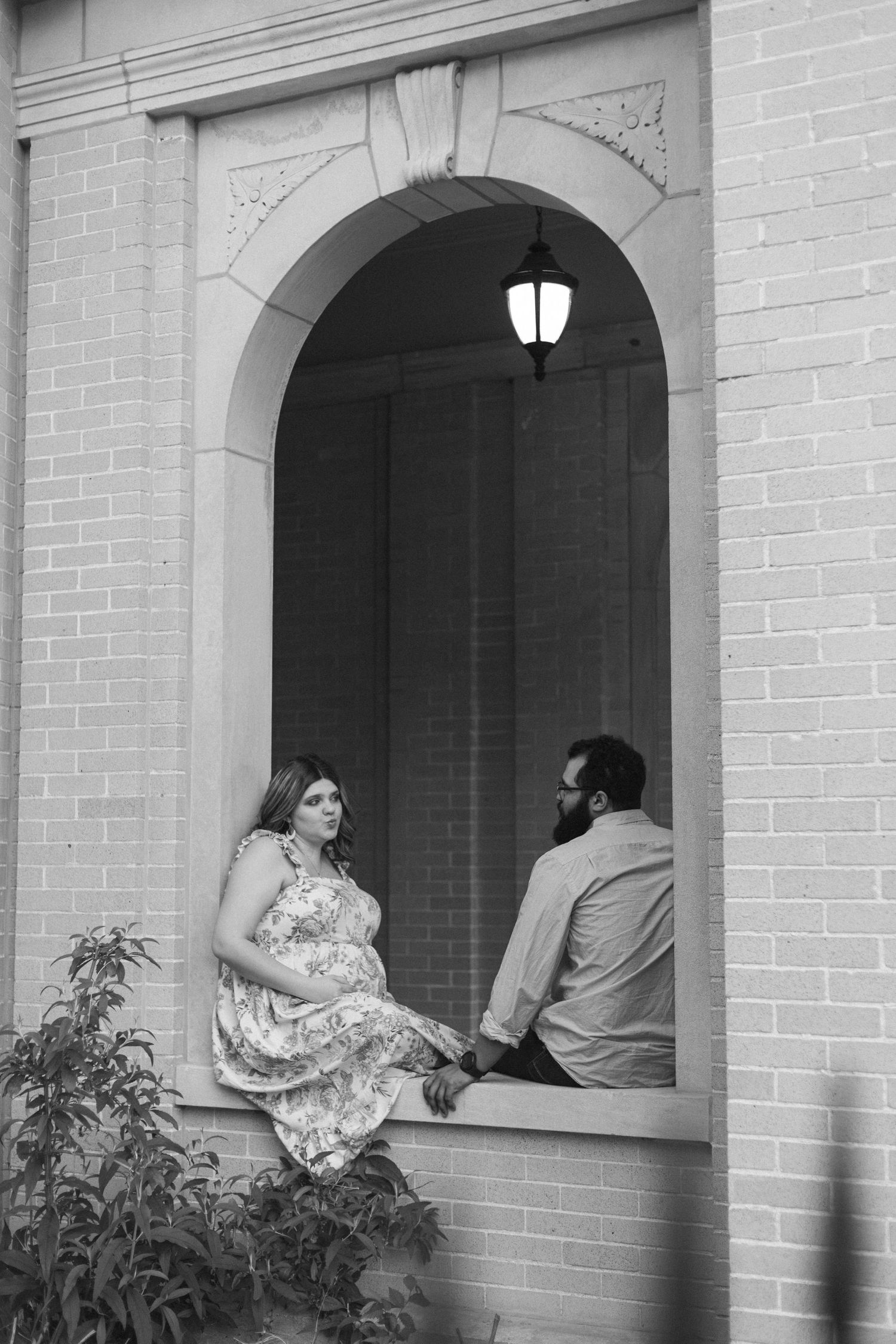 A couple poses together near arched windows of a light colored brick building in a black and white photo.
