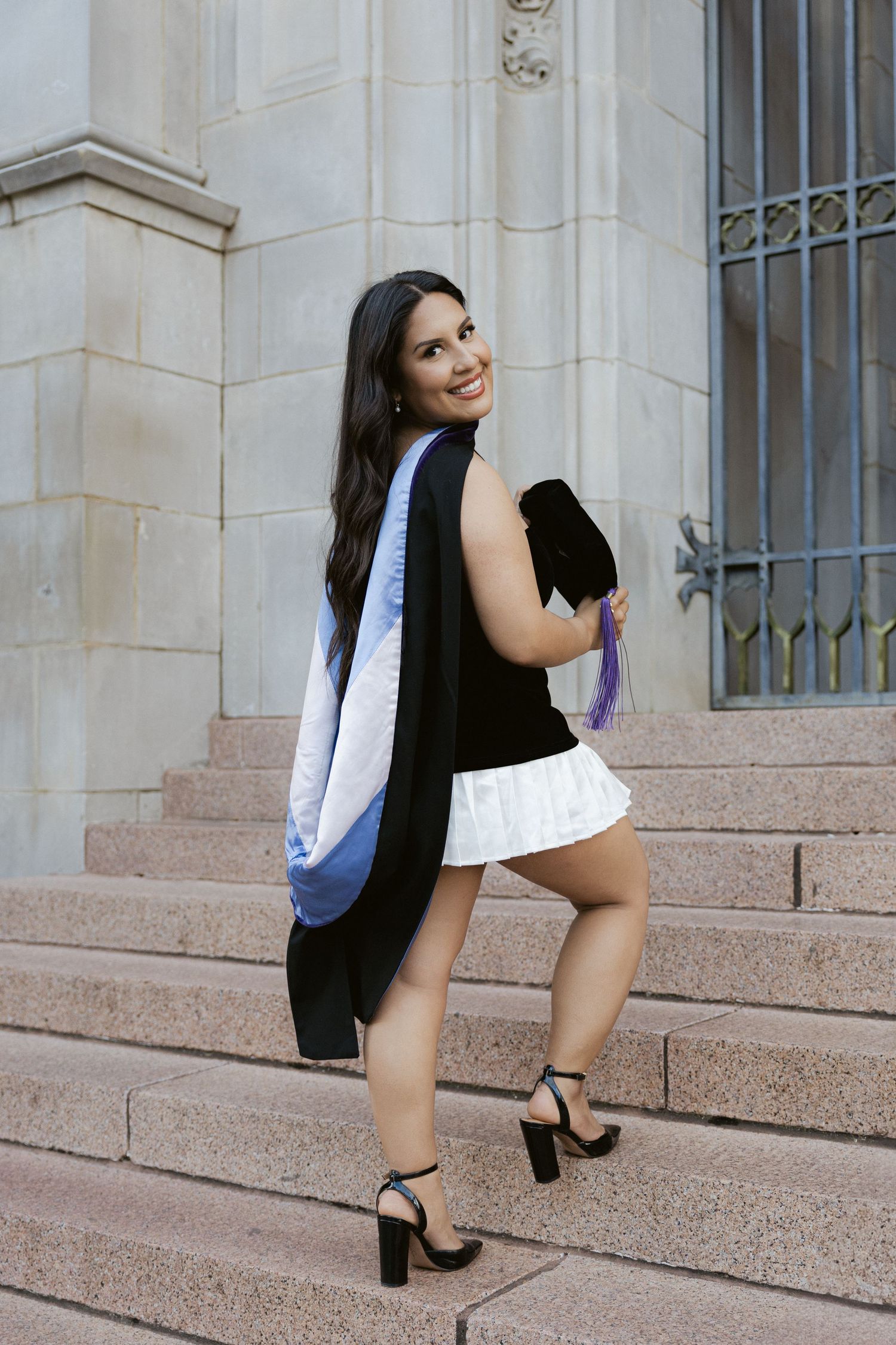 A figure in a white blazer and black skirt strikes a series of poses on steps in front of an elegant stone building entrance.