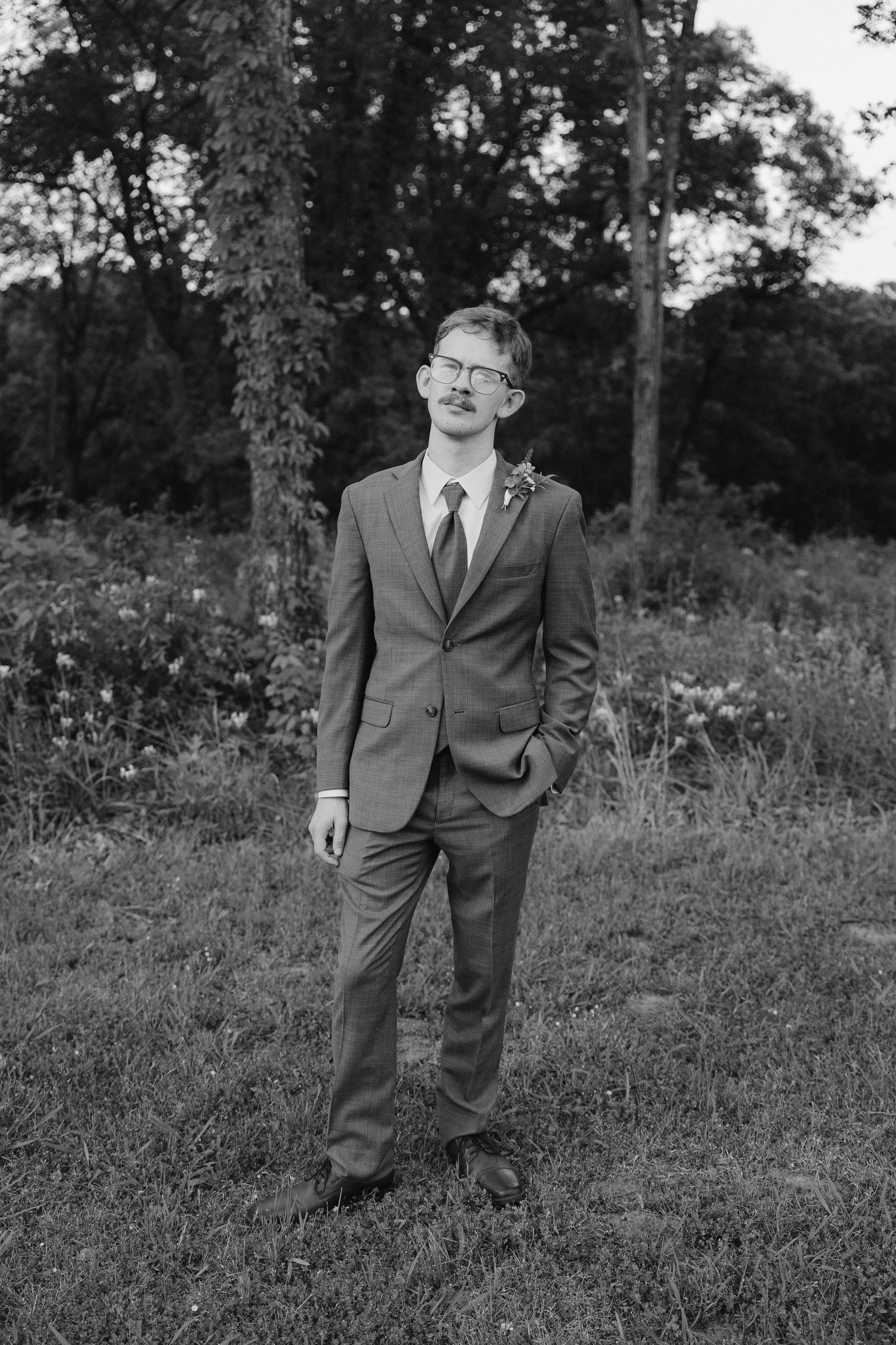 A formal portrait of a groom in a gray suit standing in a wooded outdoor setting.