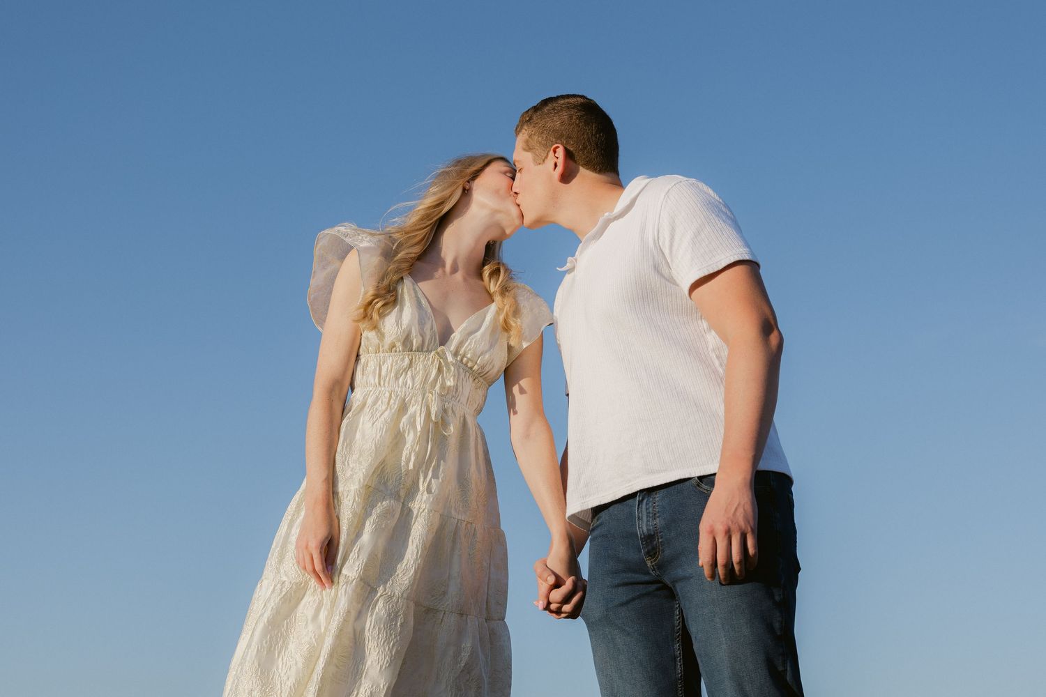 A couple stands against a bright blue sky, sharing an intimate moment during a romantic photoshoot.