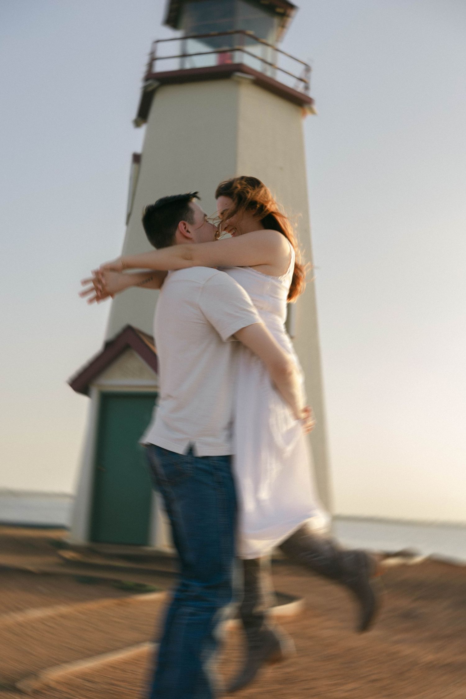 Couple sharing a romantic kiss near a lighthouse at sunset, man lifting woman in white dress.