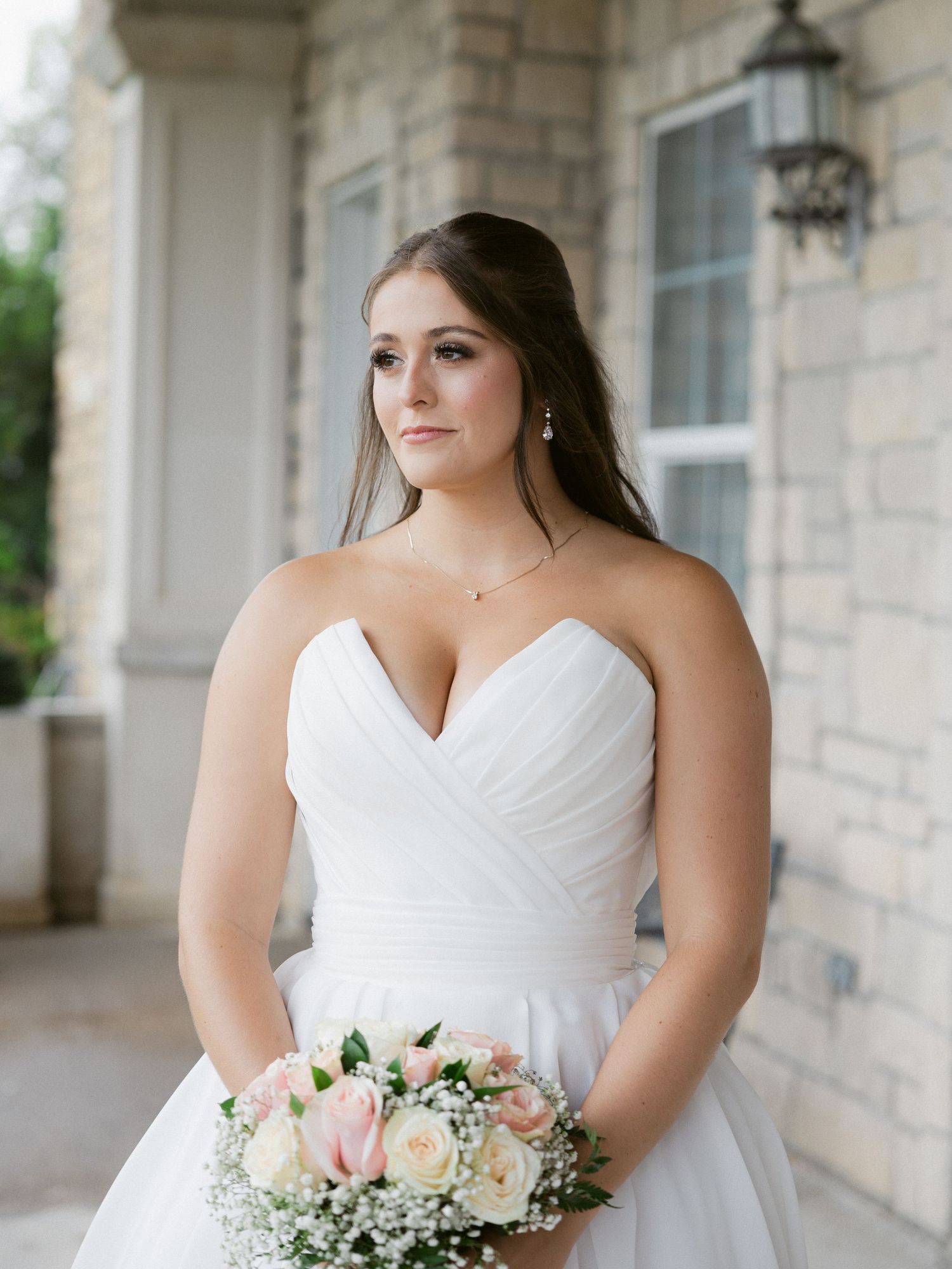 A bride in a strapless white wedding gown poses with her bouquet on a stone porch.