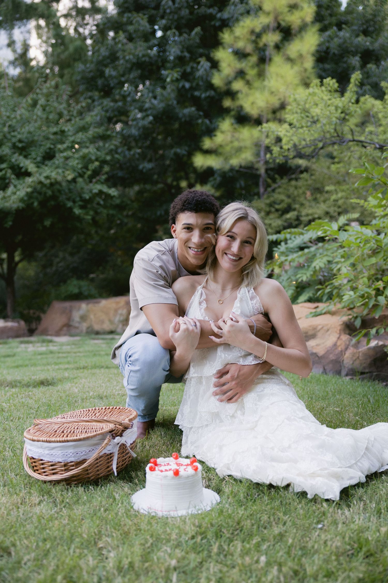 A couple enjoys a romantic picnic on the grass with a white cake and wicker basket in a garden setting.
