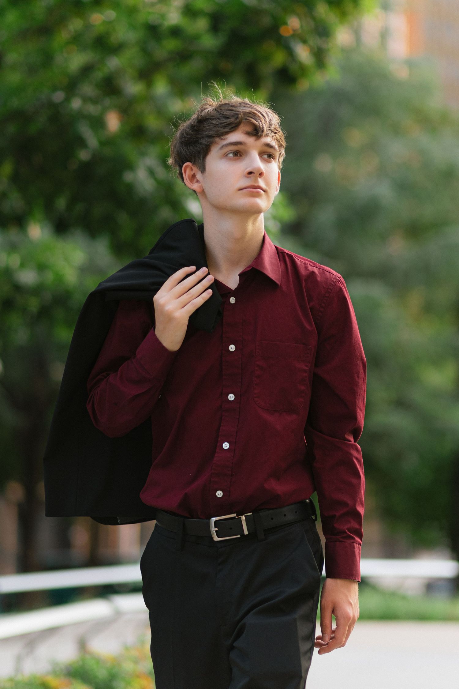 A person in a burgundy button-down shirt and dark pants poses outdoors against a blurred natural background.
