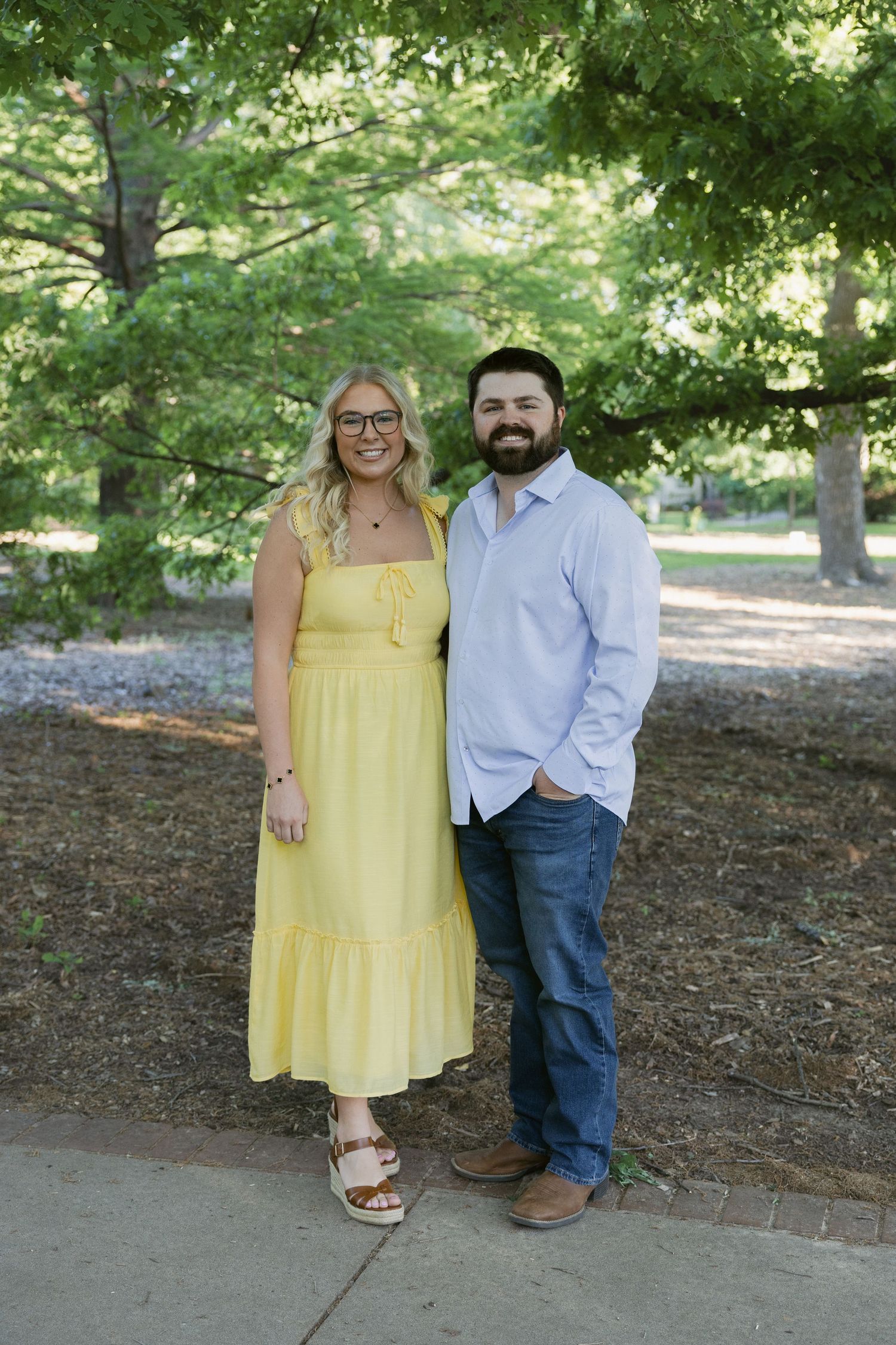A couple poses together on a path bordered by green trees, with one wearing a yellow dress and the other in blue and jeans.