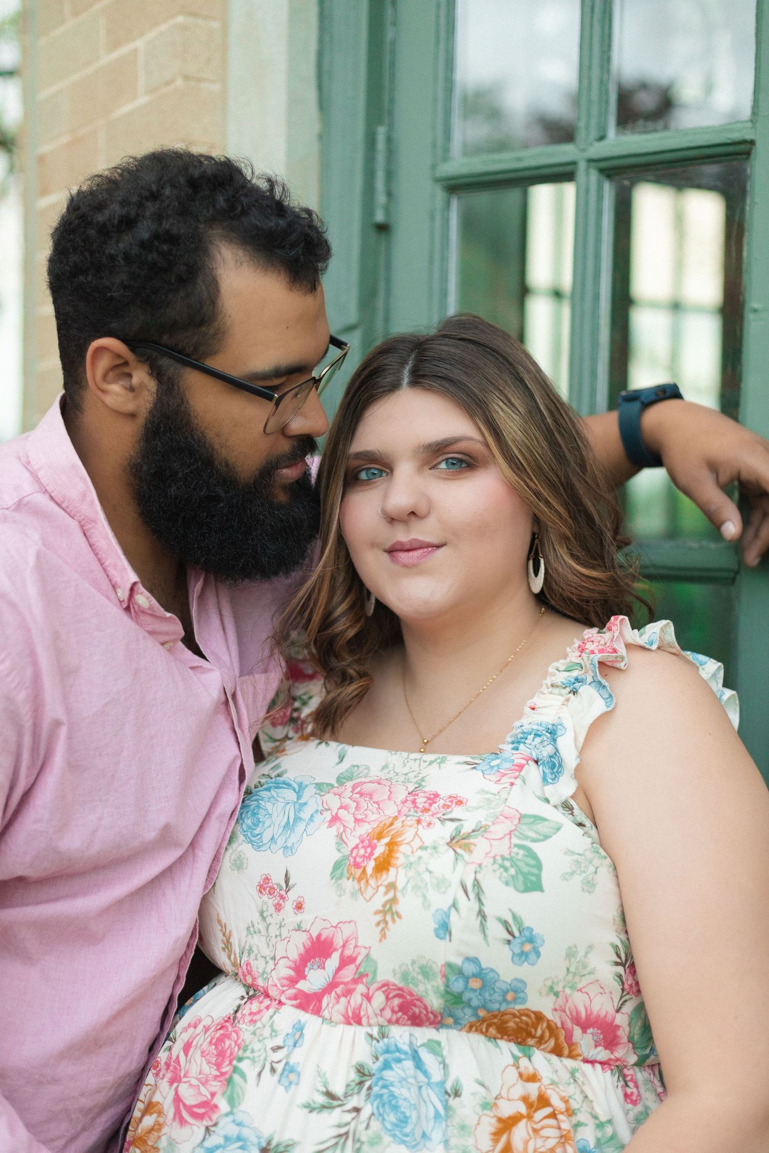 A couple shares an intimate moment outdoors by a building with green trim and windows.