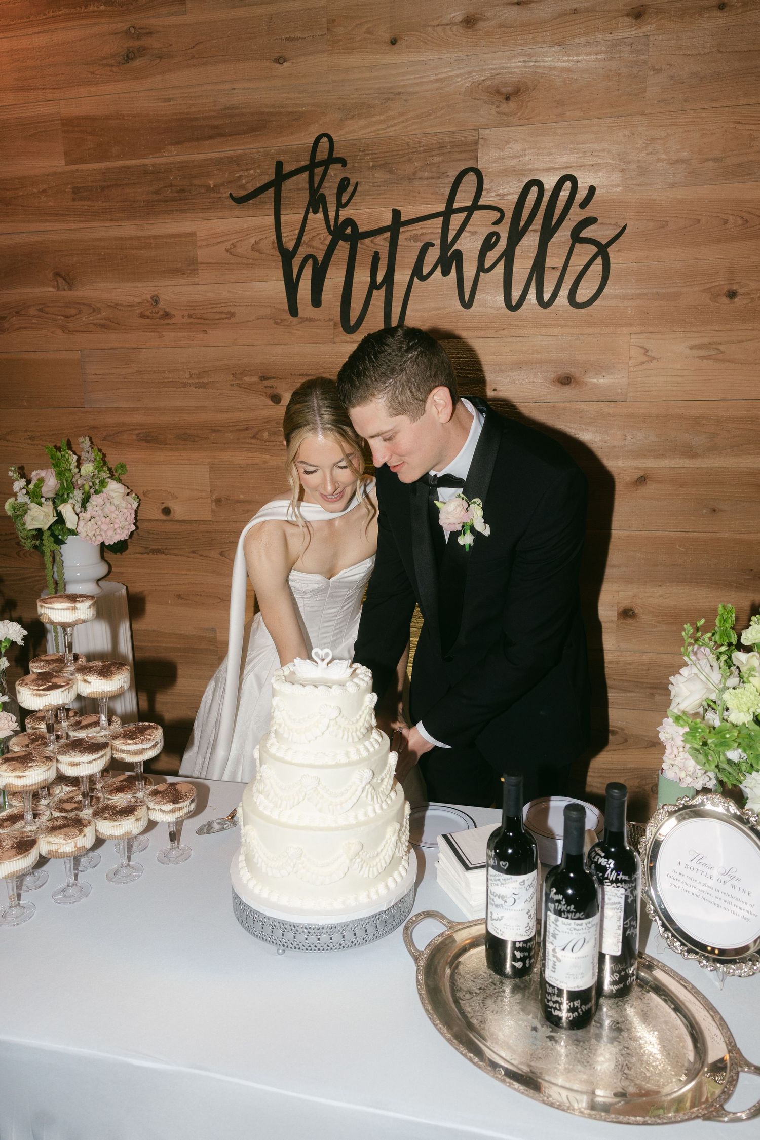 Bride and groom cutting white tiered wedding cake at reception table under The Mitchells sign.